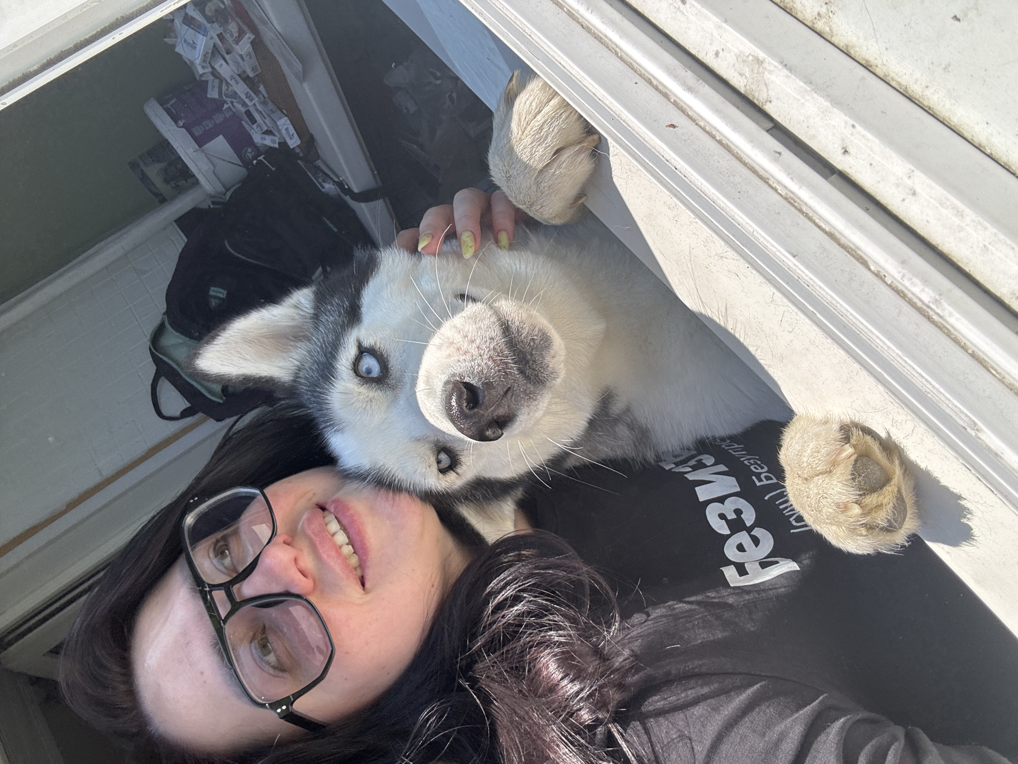 The image depicts a woman with dark hair and glasses lying down with a husky dog. The dog is leaning over her, with one paw resting on her. The background includes a window with miscellaneous objects. Location: Unknown, Unknown.