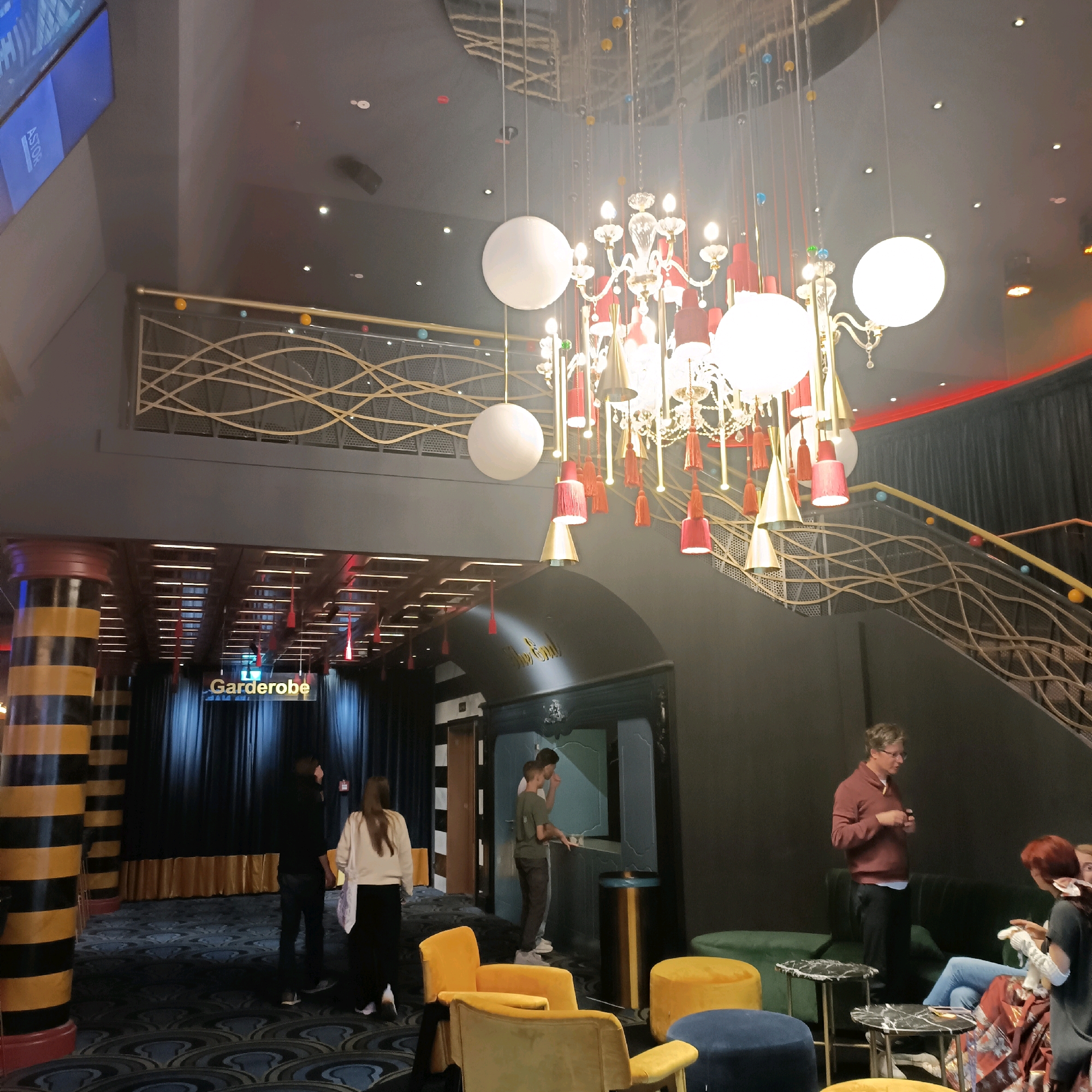 The image depicts the interior of a modern venue, possibly a cinema or theater, with a focus on its elaborate decorative elements and seating areas. In the foreground, several upholstered seats in yellow and blue are arranged in a lounge area. A man in a maroon sweater stands near a green sofa, appearing to address a woman with red hair who is seated.
In the mid-ground, a staircase with a distinctive gold, wave-like railing is visible, adorned with small colored balls. Above the staircase, a large, ornate chandelier with red and gold accents hangs, complemented by spherical white lights and dangling red and gold tassels. To the left, a tall pillar with black and yellow stripes stands prominently. A sign that reads "Garderobe" (cloakroom) is illuminated above a dark curtained area.
In the background, a large screen displaying the word "ASTOR" is mounted high on a wall. People are seen interacting in various parts of the venue, including two individuals near what appears to be a service counter or information desk. The flooring is carpeted with a patterned design in blue and grey. The overall ambiance suggests a sophisticated and comfortable gathering space. This venue is the Astor Film Lounge in Frankfurt, Germany.
