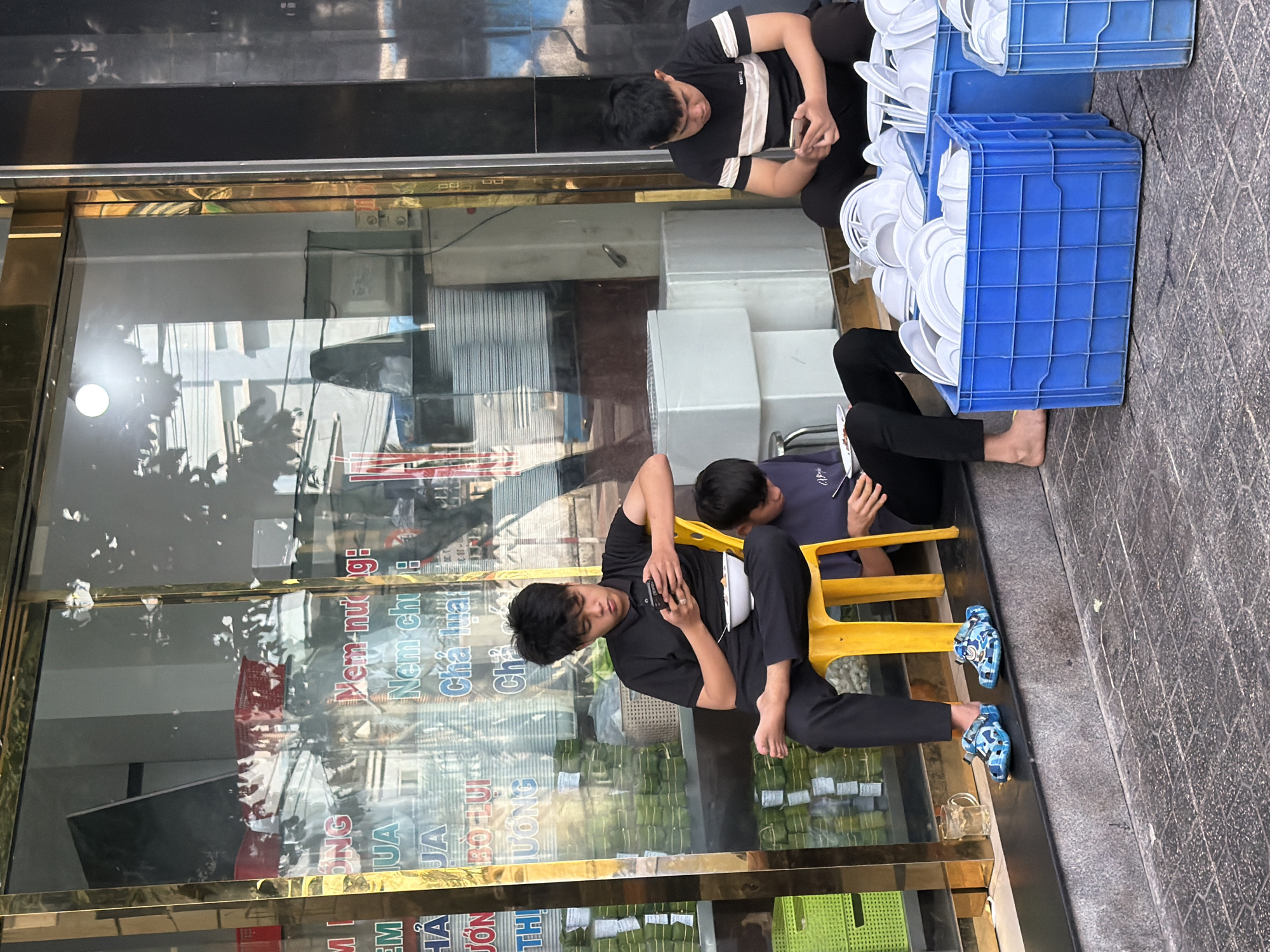 In Nha Trang Ward, Vietnam, on a sidewalk in front of a shop, three young men are seated. The man on the left, wearing a black polo shirt and black pants, sits on a yellow plastic chair. He is looking at his phone and holding a white bowl with food. He is barefoot and wearing blue camouflage clogs. To his right, a second man in a dark t-shirt and black pants is seated on the ground, also looking at his phone and holding a plate. Further to the right, a third man in a black and white striped t-shirt and black pants is seated on the ground, looking at his phone. In front of the men are several blue plastic crates filled with white plates and bowls, suggesting a food service establishment. Behind the men, the shop window displays signage, and stacks of what appear to be food items wrapped in green leaves are visible. The environment is outdoors, with a tiled sidewalk and a concrete step.
