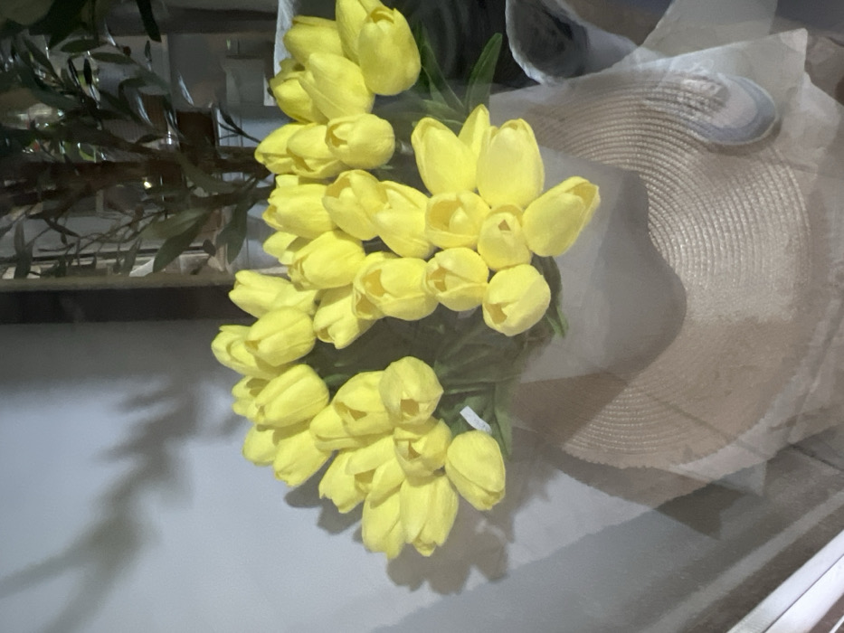 A close-up, elevated view captures a large collection of approximately 50-60 bright yellow artificial tulips, arranged in multiple bunches, resting on a light-colored, woven, circular object resembling a straw hat or mat. The tulips display closed to partially open buds with visible green stems and some green foliage. The base of the tulip arrangements is encased in a translucent, light-colored wrapping material. To the immediate left, a plain light grey wall is visible, exhibiting a distinct dark shadow, possibly from a plant or tree branch. To the right and in the background, out of focus, is a large potted plant or artificial tree with dark green leaves and a central trunk. No individuals are present. The scene is illuminated by artificial light, indicative of an indoor setting within the city of Smederevska Palanka Municipality, Serbia.