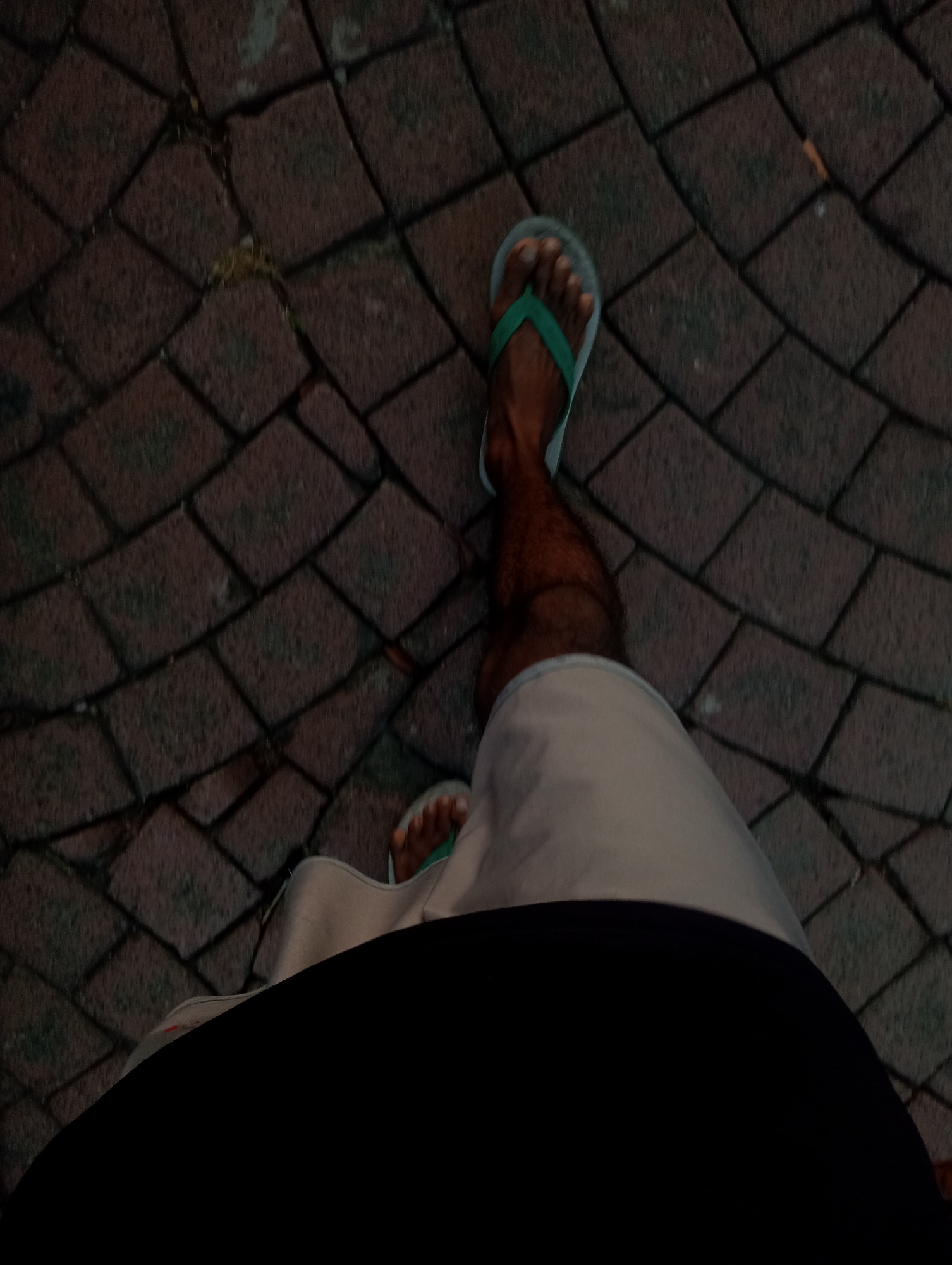 The image shows a person's legs and feet wearing light-colored shorts and green flip-flops, standing on a brick-paved walkway. The bricks are arranged in a diamond pattern and appear to be damp, suggesting it might have rained recently. There is some light foliage visible between the bricks. The perspective is from a low angle, looking down at the person's legs and the walkway. The bottom part of the image is obscured by a large black area, which could be a shadow or a part of the camera's housing. The lighting is dim, and the overall tone of the image is dark and muted. The location in Surabaya, Indonesia, is not specified in the image, but the context of brick walkways is common in many urban and public spaces there.