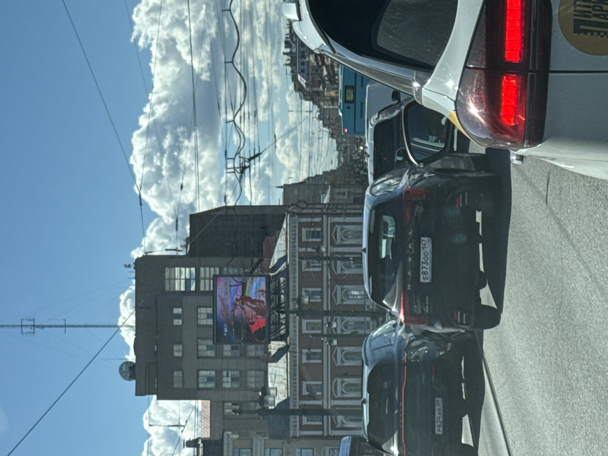 The image depicts a street scene in Saint Petersburg, Russia, taken from inside a vehicle. The view is of a city street with traffic congestion, as evidenced by the multiple cars in close proximity. A large building with a digital billboard displaying an image is visible. Overhead electrical wires are strung across the sky. The sky is blue with scattered clouds. The cars are various models, and their license plates are visible.