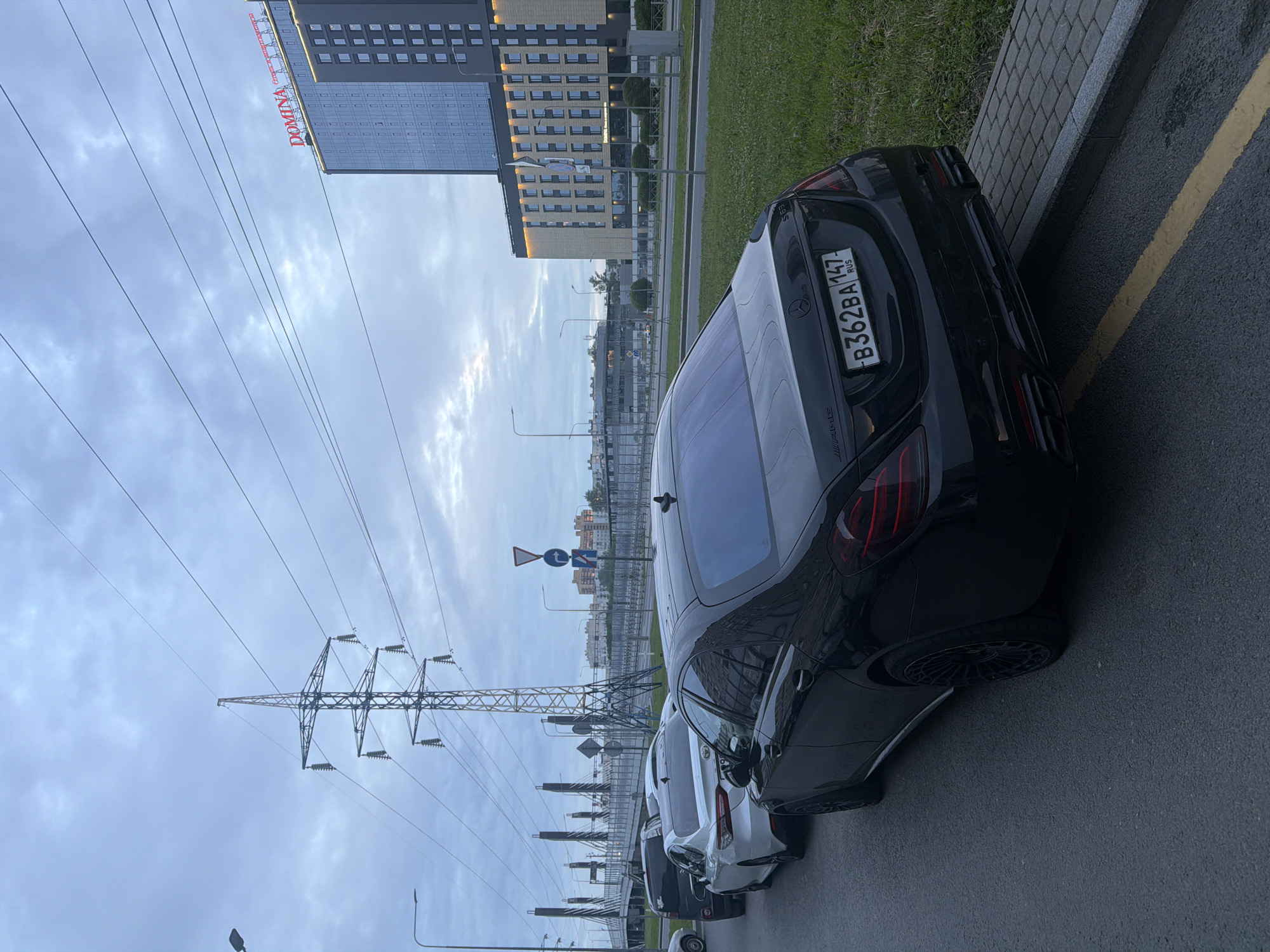 The image captures an exterior urban scene under a cloudy sky. A black Mercedes-Benz S63 AMG sedan is parked in the foreground, facing right. The car has tinted windows, black rims, and a license plate reading "B362BA 147 RUS". To the left of the Mercedes, a white Toyota Camry and another dark-colored sedan are parked in a line. In the background, a tall, modern hotel building with a "DOMINA" sign in red lettering is visible. A large electricity pylon stands prominently to the left of the vehicles, with power lines stretching across the sky. Further in the distance, more buildings and a bridge structure can be observed. The ground is paved with asphalt and the edge of the road is bordered by a sidewalk with cobblestone pavers and a strip of grass.