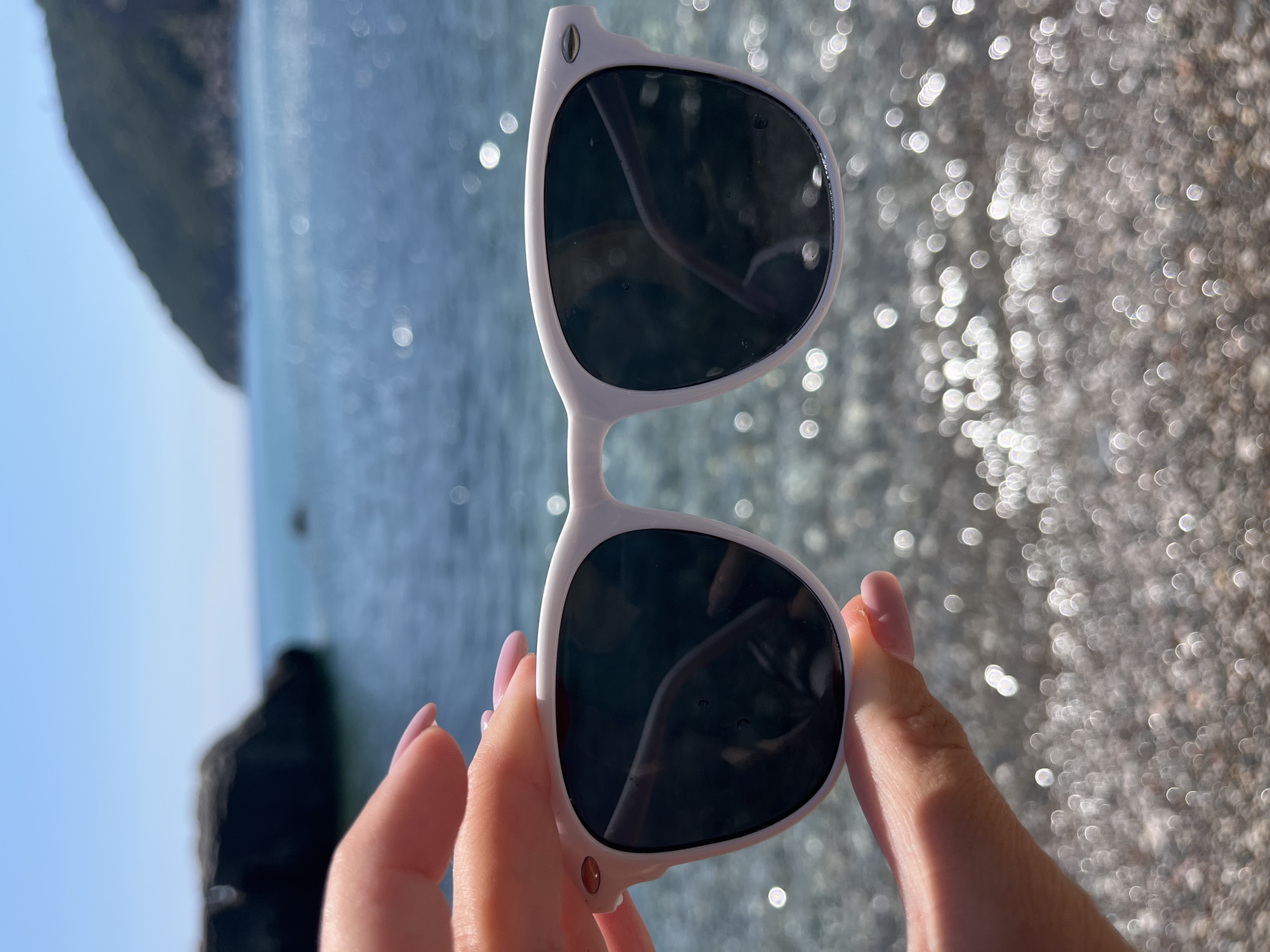 A pair of white sunglasses with dark lenses is held up by a person's hands. The sunglasses are in the foreground, and the background is out of focus, showing a body of water and a pebbly shore. The water is blue and appears calm, with sunlight reflecting off its surface. The shore is made up of small, light-colored pebbles. The scene suggests a sunny day, likely at a beach or seaside location. The person's hands have light pink painted fingernails and are holding the sunglasses by the temples. The overall mood is relaxed and summery. The location is identified as Zagrađe, Montenegro.