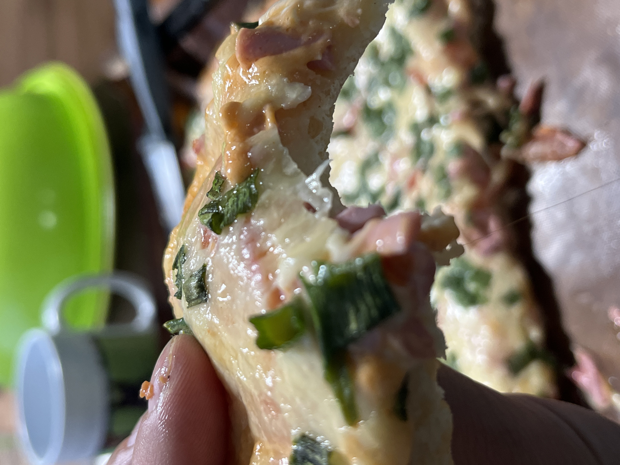 A close-up, slightly blurred shot focuses on a slice of pizza being held by a person's thumb and forefinger. The pizza appears to have a golden crust, melted cheese, small pieces of ham, and green herbs, possibly chives or parsley. A bite has been taken out of the slice, revealing the texture of the dough and toppings. In the background, out of focus, is a bright green object that could be a bowl or container, and a white lid or cap. Further back, a dark object, possibly a kitchen utensil, is also visible. The lighting suggests an indoor setting, possibly a kitchen. The overall impression is casual and domestic, focusing on the enjoyment of food. There is no visible text in the image.
