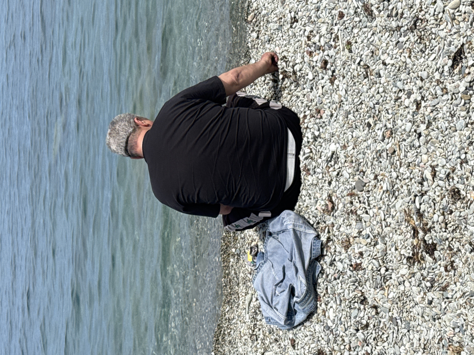 A person is sitting on a rocky beach next to the water. The person is wearing a black shirt, and a denim jacket is lying next to them on the ground. The water is a light blue color, and the rocks on the beach are small and white. The city is unknown, and the location is unknown.