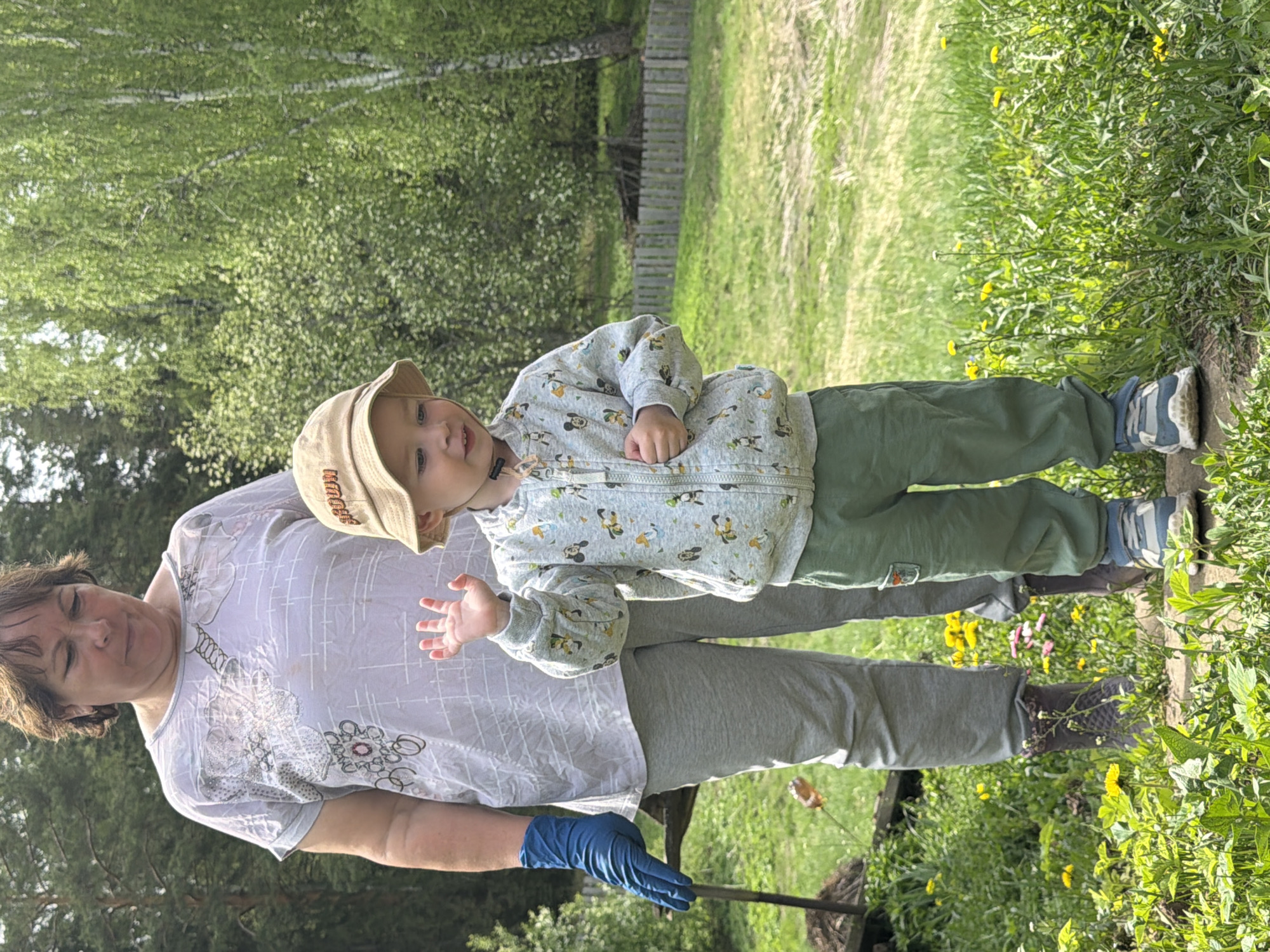 The image depicts a woman and a child standing outdoors. The woman is wearing a light-colored t-shirt with floral patterns, grey pants, and blue gloves. She is holding a tool in her gloved hand. The child is wearing a light-colored zipper jacket with Mickey Mouse patterns, green pants, a beige hat, and blue and grey shoes. They are standing in a grassy area with dandelions, near a wooden fence and trees. The location is identified as Уральское, Russia, based on the prompt. The photo is rotated to the left.