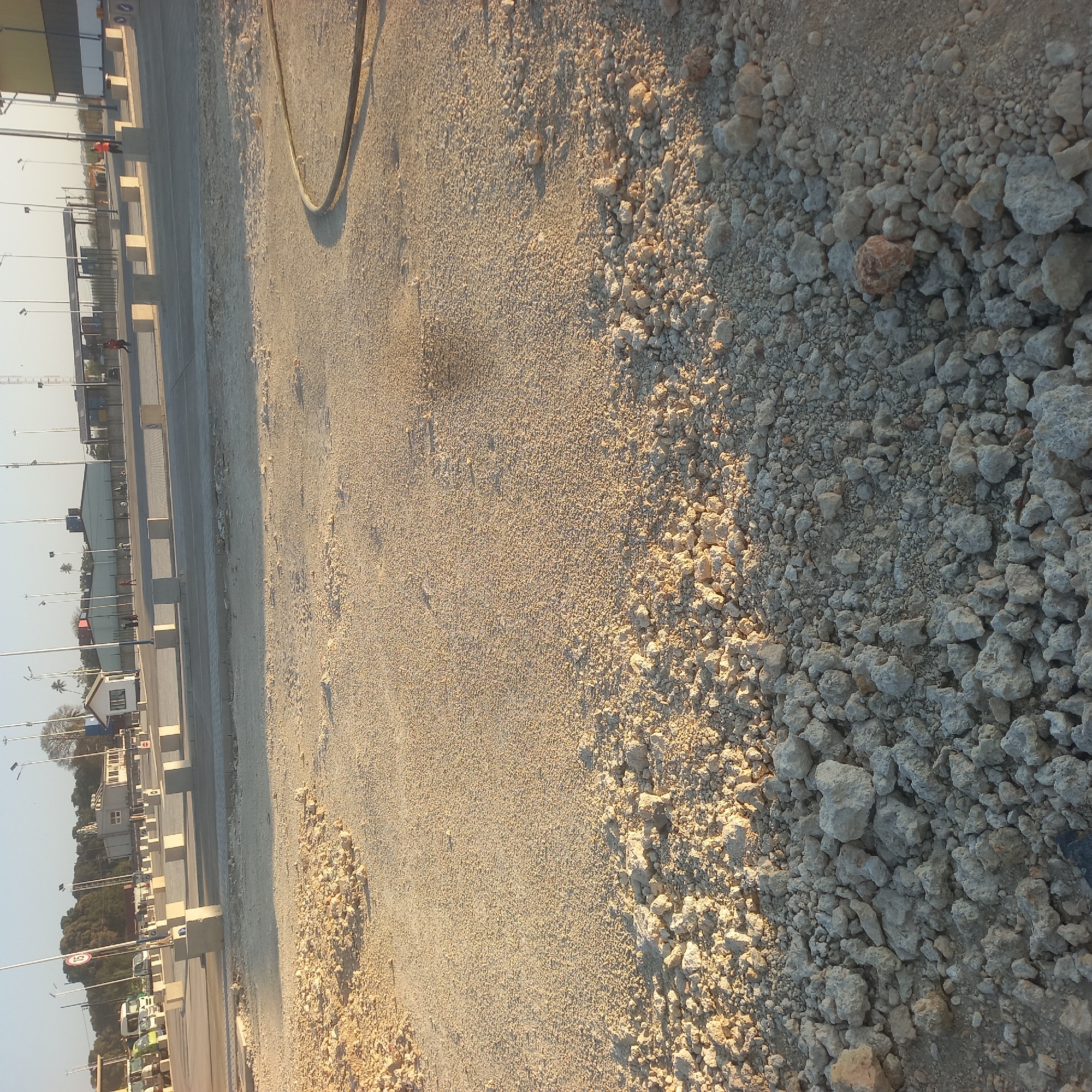 The image shows an outdoor construction site. The ground is covered in gravel and some larger rocks, with piles of rubble in the foreground. In the background, there are several buildings and structures, including a small white building with a red roof, and a longer, low-rise building with shipping containers visible. There are also several streetlights and some vehicles, including trucks and what appear to be service vehicles. Several people are visible in the background, some wearing orange vests, indicating they are likely workers. The scene appears to be an area undergoing development or renovation. The presence of shipping containers and the overall aesthetic suggests a port or industrial area. The location is Bububu, Tanzania.