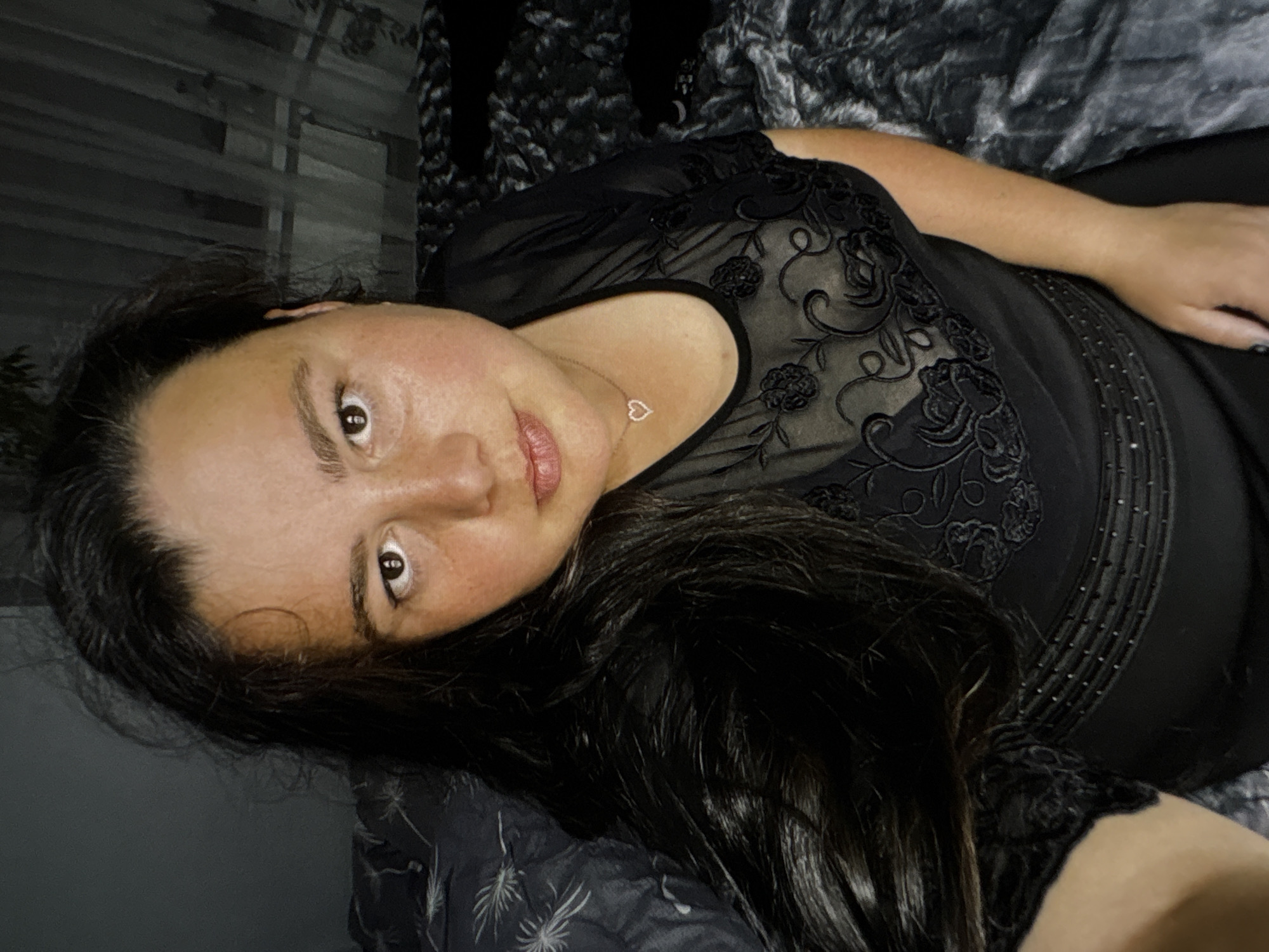 A medium shot shows a woman from the chest up, resting on a bed with dark bedding. She has long, dark, wavy hair that falls over her left shoulder and chest. Her skin is fair with freckles. She is wearing a black sheer top with floral embroidery. A delicate heart-shaped necklace with a small gem is visible on her chest. The background is dimly lit, with curtains visible on the right. The woman's gaze is directed towards the camera.