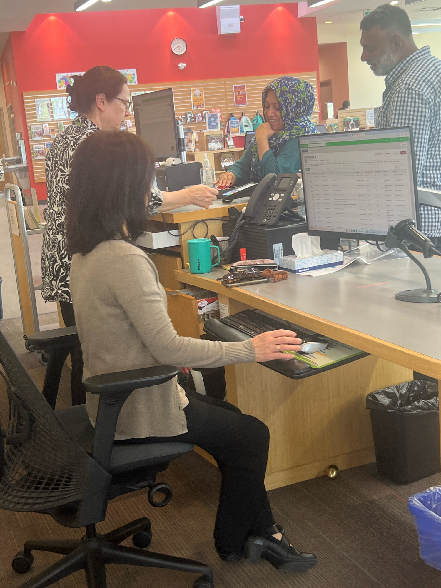 The image depicts an indoor scene, likely a library or a similar public service desk, with four individuals visible. In the foreground, a woman with dark hair is seated at a desk, facing away from the camera, interacting with a computer. She is wearing a light-colored cardigan and dark pants. To her left, another woman with dark hair and glasses, wearing a patterned black and white top, stands and appears to be engaged in a conversation or assisting the seated woman.
Behind the desk, a woman wearing a blue and green hijab is seated and smiling, looking to her left, towards the standing woman. She is wearing a teal-colored garment. To her right and slightly behind her, a man with a beard, wearing a plaid shirt, is standing and looking at the woman in the hijab. His interaction suggests a conversation.
The environment includes a desk with computers, a telephone, a scanner, a box of tissues, and a teal mug. Shelving with books and decorative items is visible in the background, along with a clock on a red wall. The flooring is wood-patterned. The presence of multiple individuals at a service desk suggests a public space. The mention of the city of Vaughan, Canada, is not supported by any visual cues within the image itself.