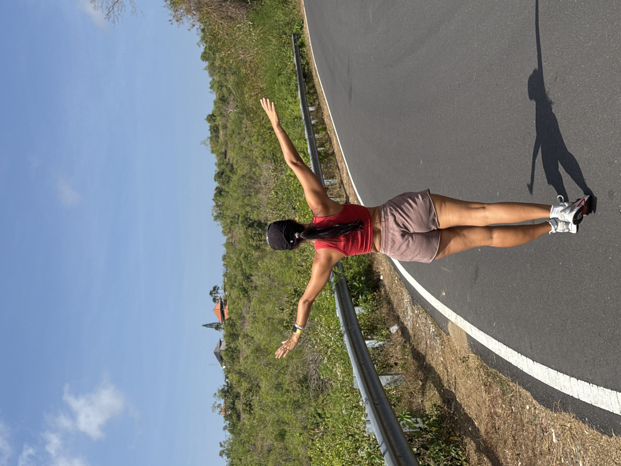 A woman is pictured from behind, walking on a paved road with her arms outstretched. She is wearing a red crop top and light brown shorts. The road curves to the right and is bordered by a metal guardrail on the left. Beyond the guardrail, there is dry grass and vegetation. In the background, a lush green hillside is covered with trees and bushes, with a few houses visible at the top. The sky is clear and blue with a few wispy clouds. The sun is shining, casting a shadow of the woman on the road. This scene is consistent with a location in Kuta Selatan, Indonesia, given the tropical vegetation and architecture.