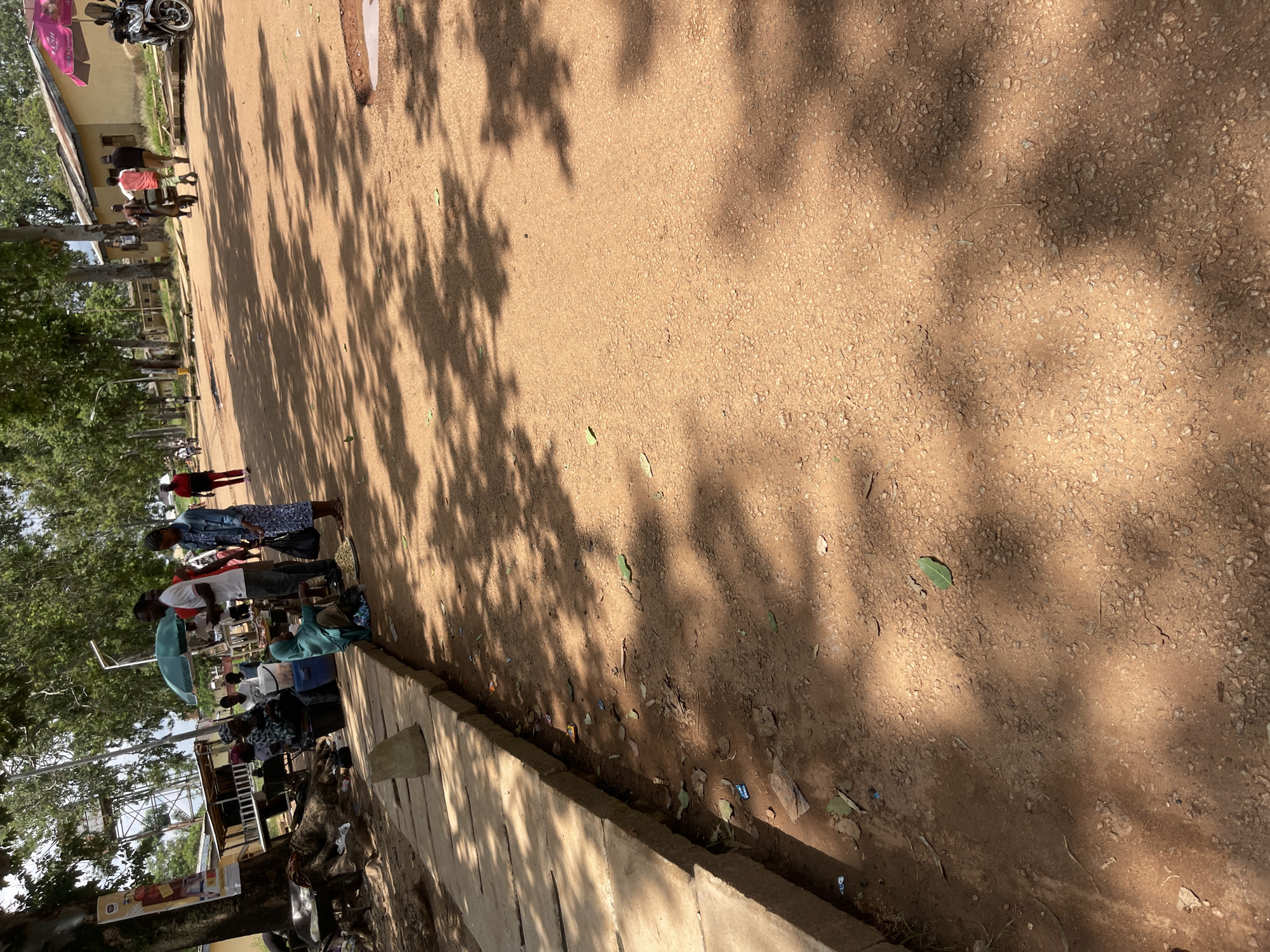 This is an outdoor scene on a sunny day, likely in the city of Rafin Par, Nigeria, showing a dirt road with scattered small rocks and fallen leaves. The ground is partially covered by dappled sunlight and tree shadows. On the left, there is a concrete sidewalk or raised platform with several people gathered around. Some are sitting, and others appear to be selling goods. One person is holding an umbrella. Further down the road, other individuals are walking or standing, and there's a glimpse of buildings with yellow walls and a pink umbrella in the background. A motorcycle is parked near the buildings.