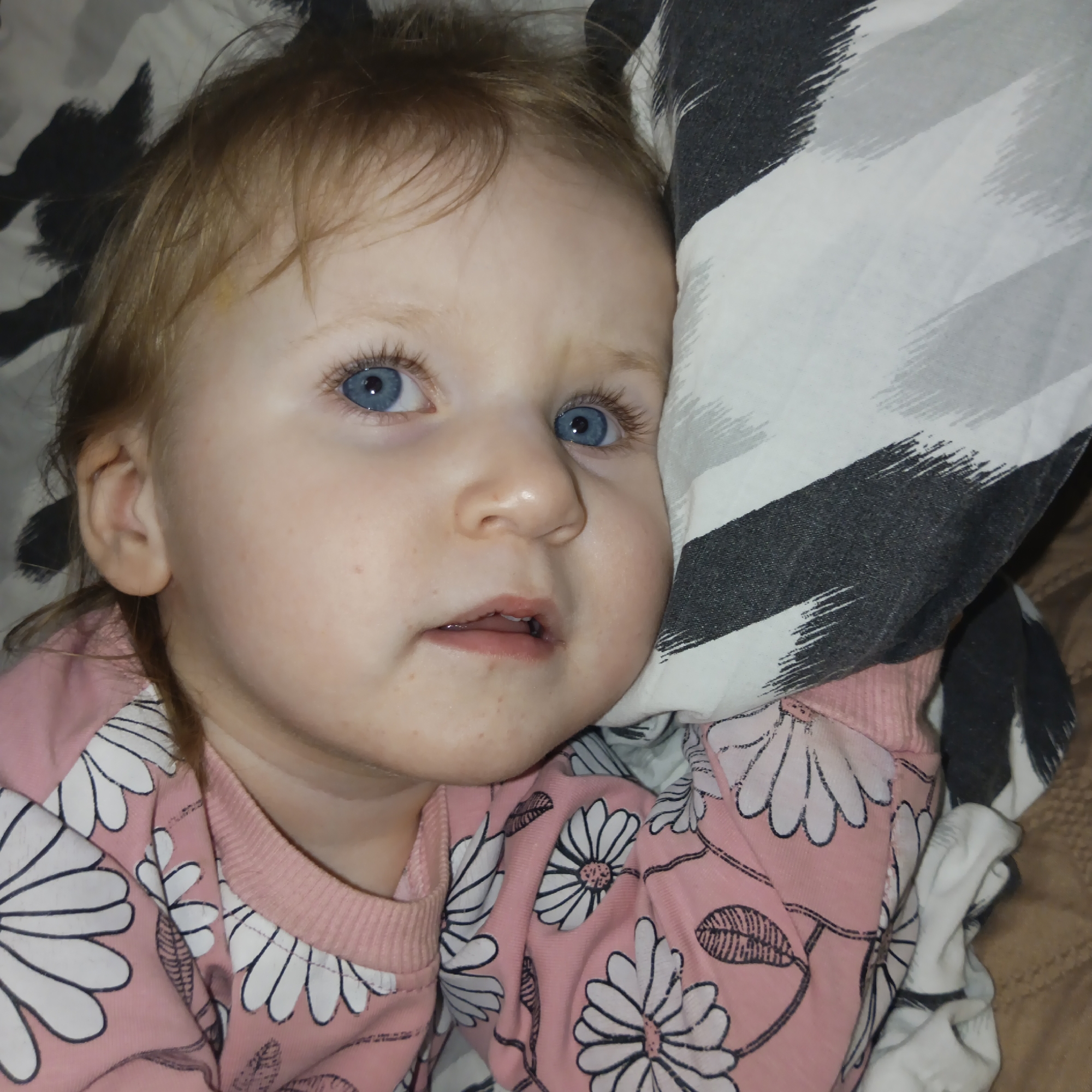 A young child with fair skin and blue eyes lies on a bed with a patterned pillow next to their head. The child is wearing a pink long-sleeved shirt with a white floral design. The child appears to be looking upwards. The location is not discernible from the image.