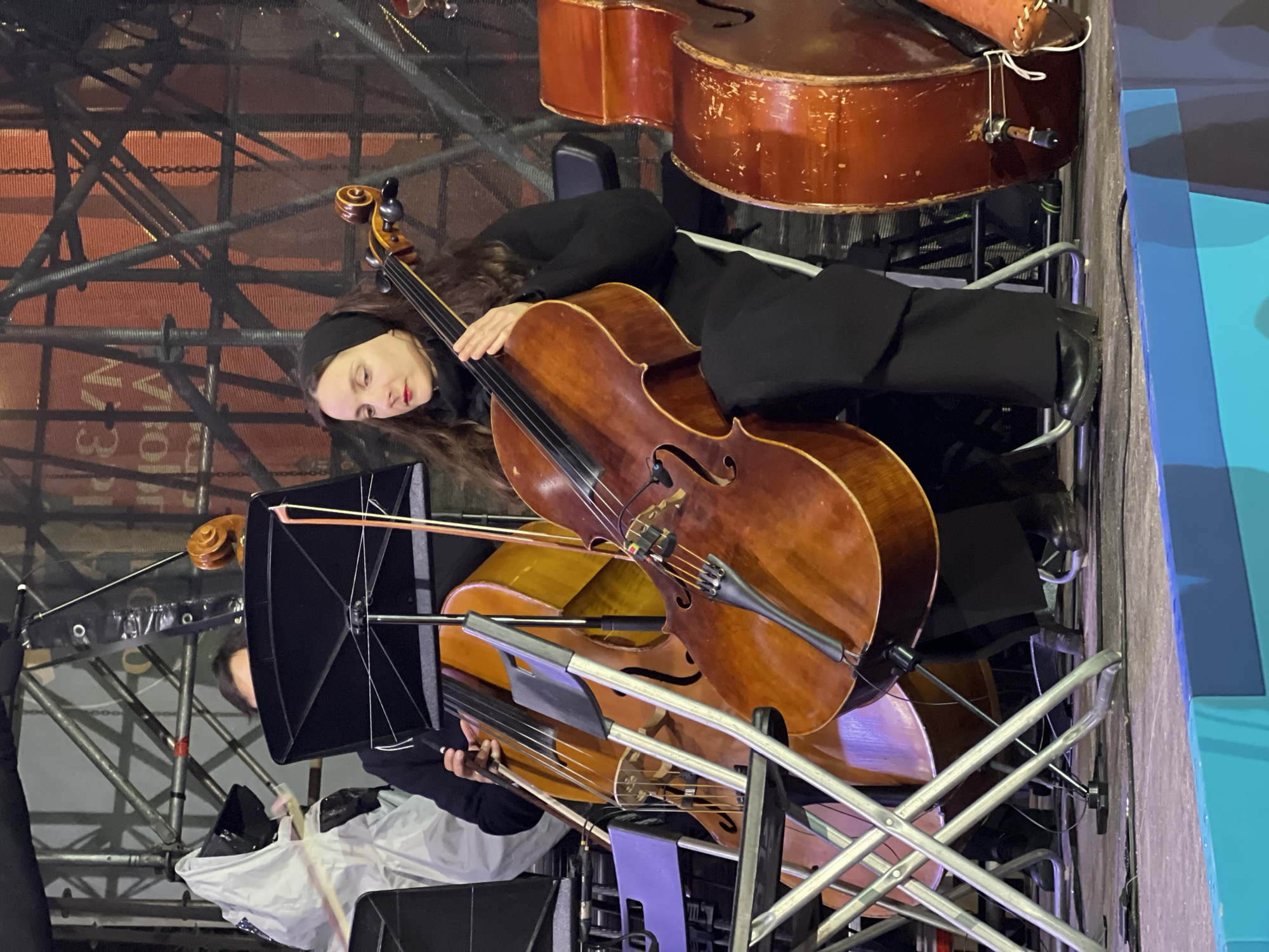 A woman with long dark hair and a black headband is seen reclining with her cheek resting on a cello. She is wearing black clothing. Two other cellos are visible, one stacked on top of the other and positioned above the woman. There is a person behind a music stand with a cello bow, and scaffolding is visible in the background. Additional musical instruments are also nearby. A section of a building or stage set is seen on the right side of the frame, with a blue color dominating the edge. The image was taken in Saint Petersburg, Russia.