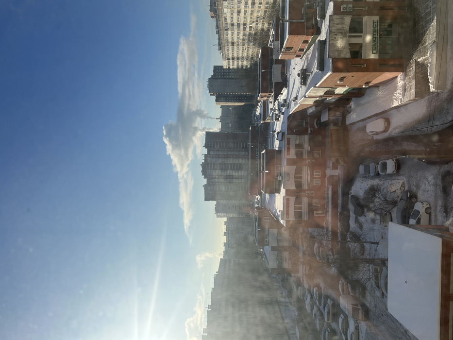 The image displays an elevated view of a snow-covered urban environment in Saint Petersburg, Russia, under a clear, bright blue winter sky. The sun, positioned in the upper left, casts a strong glare and long shadows.
In the foreground and midground, a complex of low-rise, reddish-brown brick commercial or mixed-use buildings with flat, snow-covered roofs is visible. One such building, prominently located on the right, features a large sign in Cyrillic, "ГЕМОТЕСТ" (GEMOTEST), indicating a medical laboratory. This building also exhibits a grey panel on its facade, multiple windows, and exterior air conditioning units. Snow covers the building roofs, ledges, and the ground surrounding them.
Below the foreground buildings, a network of snow-covered roads and parking lots is observed. Numerous vehicles, mostly cars, are parked, many partially or entirely covered in snow. Tire tracks are visible on the roads, indicating recent vehicular activity. Sparse, bare deciduous trees are interspersed within the snowy ground areas.
In the background, a dense array of multi-story residential apartment blocks, typical of Soviet or post-Soviet era architecture, dominates the skyline. These buildings vary in color from dark grey to lighter tones. In the far distance, to the right of center, plumes of white smoke or steam rise from industrial chimneys, forming distinct cloud-like formations against the blue sky.
No individuals or explicit human actions are visible within the frame. The scene portrays a cold, sunny winter day in a developed residential and commercial district of Saint Petersburg.