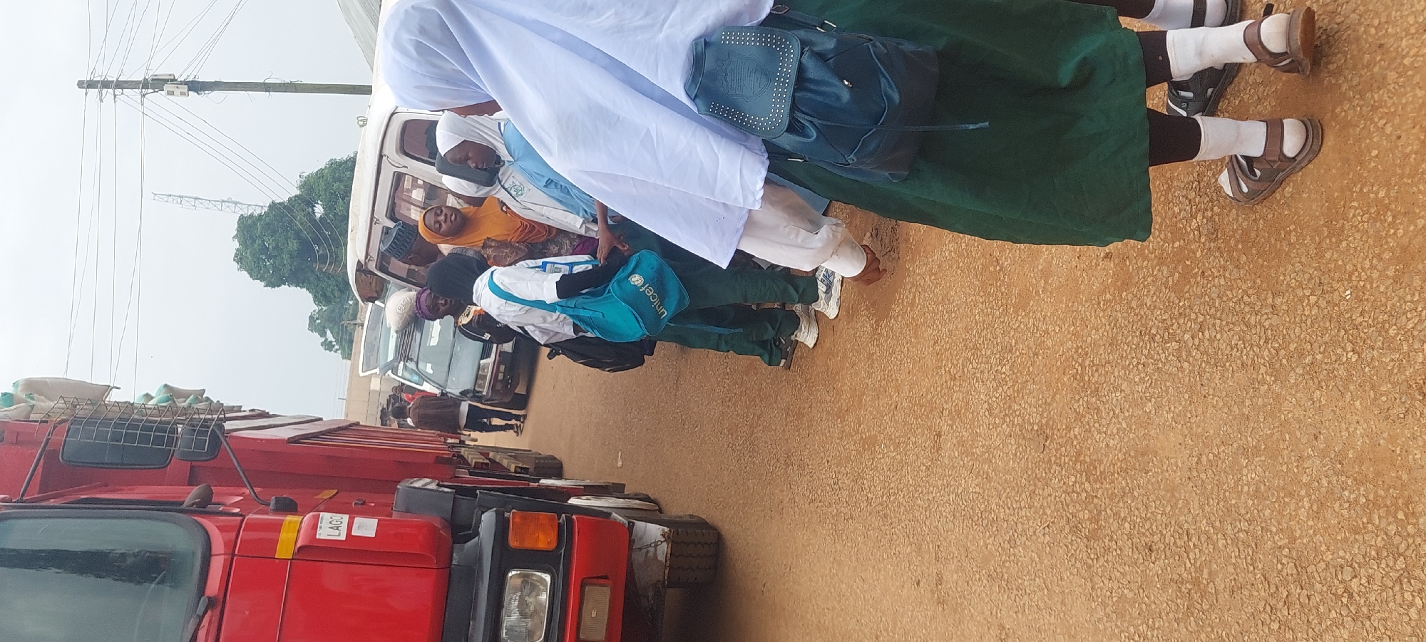 A group of people, primarily young students wearing uniforms and headscarves, stand on a dirt road next to a large red truck. The truck is loaded with sacks, and its side has a "LAGOS" sticker. In the background, several vehicles, including a white van and a dark-colored car, are parked or stopped. Power lines and a tall tower are visible in the distance. The overall scene appears to be an outdoor roadside gathering.
