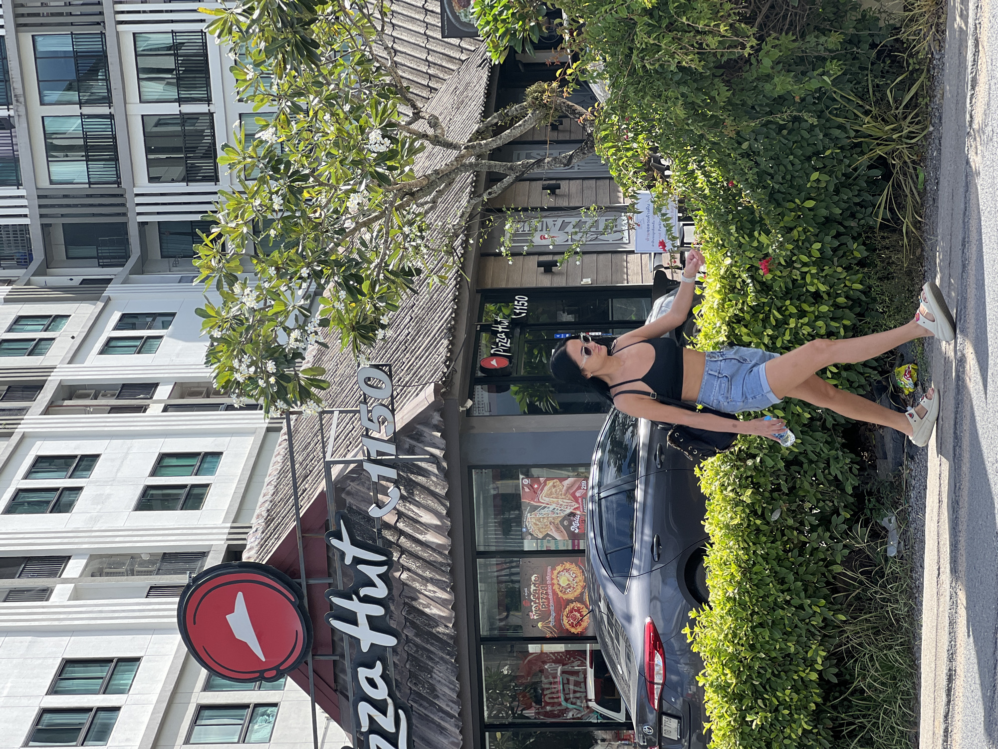 An exterior shot captures a Pizza Hut restaurant with its prominent red and white logo. The building features a textured, aged roof and a glass storefront displaying menu posters. In front of the restaurant, a dark gray sedan is parked. A woman, wearing a black crop top, denim shorts, and white sandals, walks from right to left in front of a lush green hedge. She carries a water bottle in her right hand. Behind the restaurant, a modern apartment building with multiple balconies and windows is visible. The scene is illuminated by bright daylight, casting shadows and highlighting the various architectural elements and natural foliage.