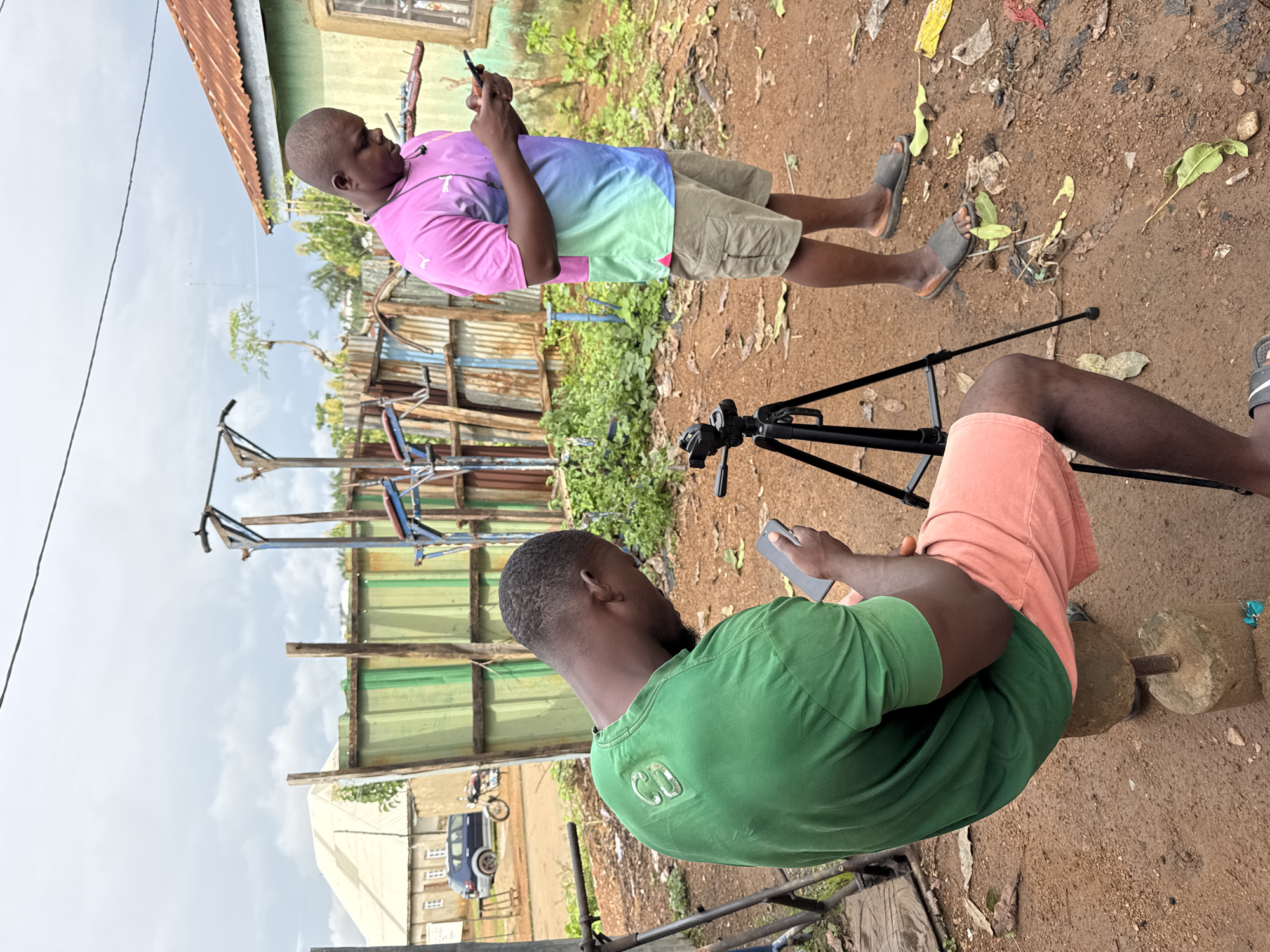 In Akwanga, Nigeria, two Black men are outdoors, focusing on their mobile phones. One man, wearing a green t-shirt and peach shorts, is seated on a makeshift stool constructed from concrete blocks and a metal rod. He holds a smartphone in his right hand, his head bowed in concentration. To his right, a black tripod stands with a camera mount. The second man, dressed in a vibrant pink, blue, and green t-shirt and beige cargo shorts, stands with his phone held in his hands, also engrossed in its screen. His left ear is adorned with a black earphone.
The environment is rustic. A corrugated metal fence, showing signs of rust and age, forms a backdrop. Beyond the fence, a light-colored building with a peaked roof and windows is visible, as is a dark blue car parked on a dirt road. Several trees and overgrown green foliage are scattered throughout the scene. The ground is a mixture of dirt and scattered debris, including leaves and plastic waste. The sky above is overcast with grey clouds. The overall impression is one of a casual, possibly informal, setting.
