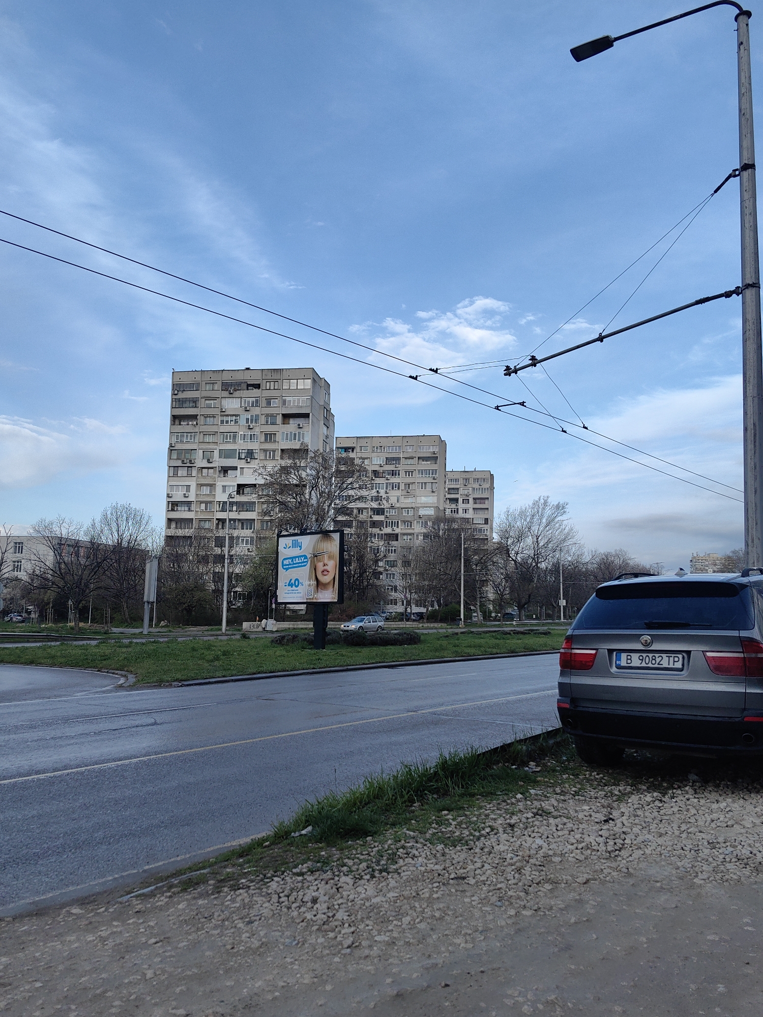 The image captures a street scene in Varna, Bulgaria, featuring a gray BMW SUV parked on a gravel shoulder next to a wet, asphalt road. The license plate of the car is visible as "B 9082 TP." In the background, several large, prefabricated apartment buildings dominate the view, typical of Soviet-era construction. A billboard advertising a hair product with a blonde woman's face is positioned in front of the buildings. Bare trees line the green median. Overhead trolley wires and a streetlight extend above the frame. The sky is overcast with patches of blue.