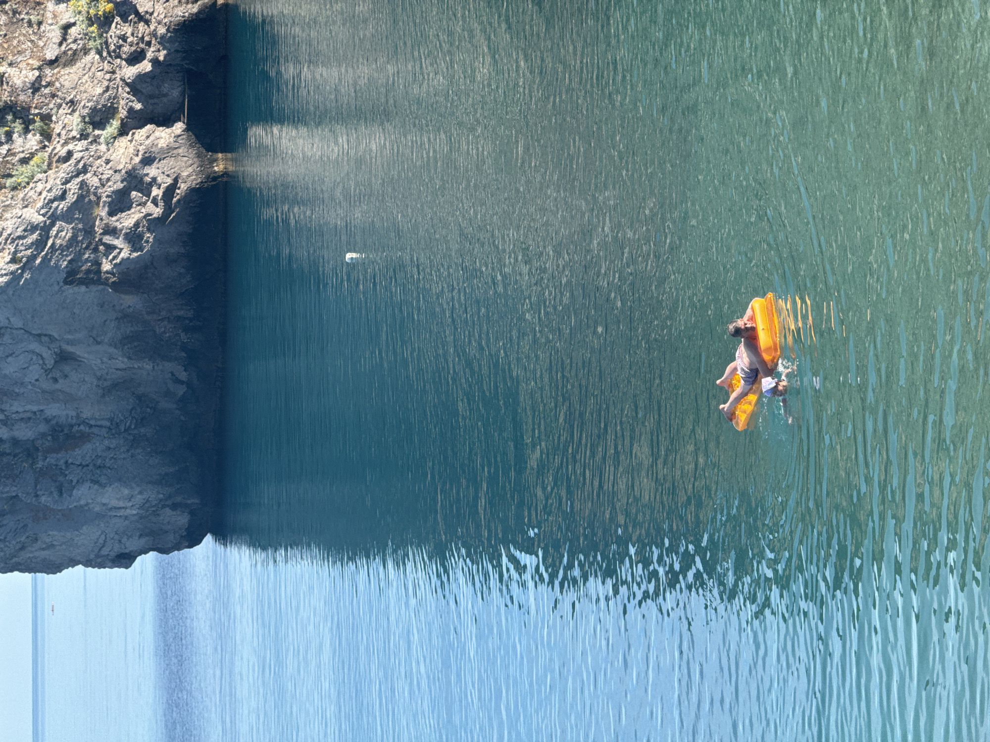 The image depicts a person relaxing on an inflatable raft in a body of water, likely a lake or sea. The person, wearing striped shorts, is lying on their back on a bright orange inflatable raft. The water is a clear, deep blue-green and appears calm with gentle ripples reflecting the sunlight. To the left of the frame, a rocky cliff face rises from the water, covered in sparse vegetation. The sunlight suggests it is daytime, with clear weather. The scene conveys a sense of tranquility and leisure, with the person enjoying a peaceful moment on the water. The reflection of the cliff and the sky on the water creates a striking visual contrast between the solid landmass and the fluid surface.
