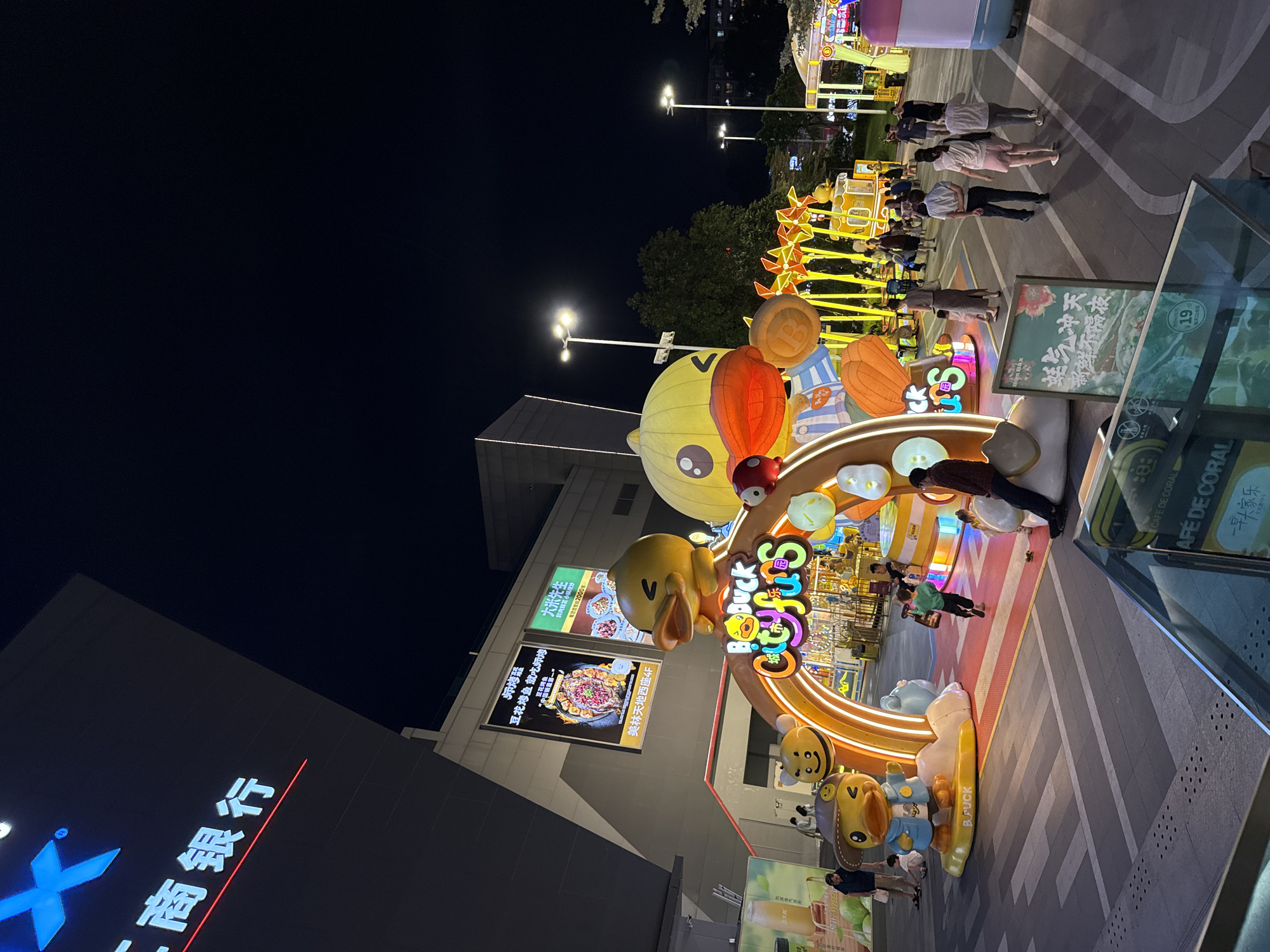 This is a nighttime, eye-level, medium shot of a plaza outside a building in Tianhe District, China. A large, brightly lit archway featuring cartoon ducks is the central focus, with the words "Big Duck City funs" prominently displayed. Several people, including families and individuals, are present, walking through the plaza and interacting with the decorations. A building with illuminated signage is visible in the background. Streetlights illuminate the area, and the sky is dark. The ground is paved with a patterned surface.