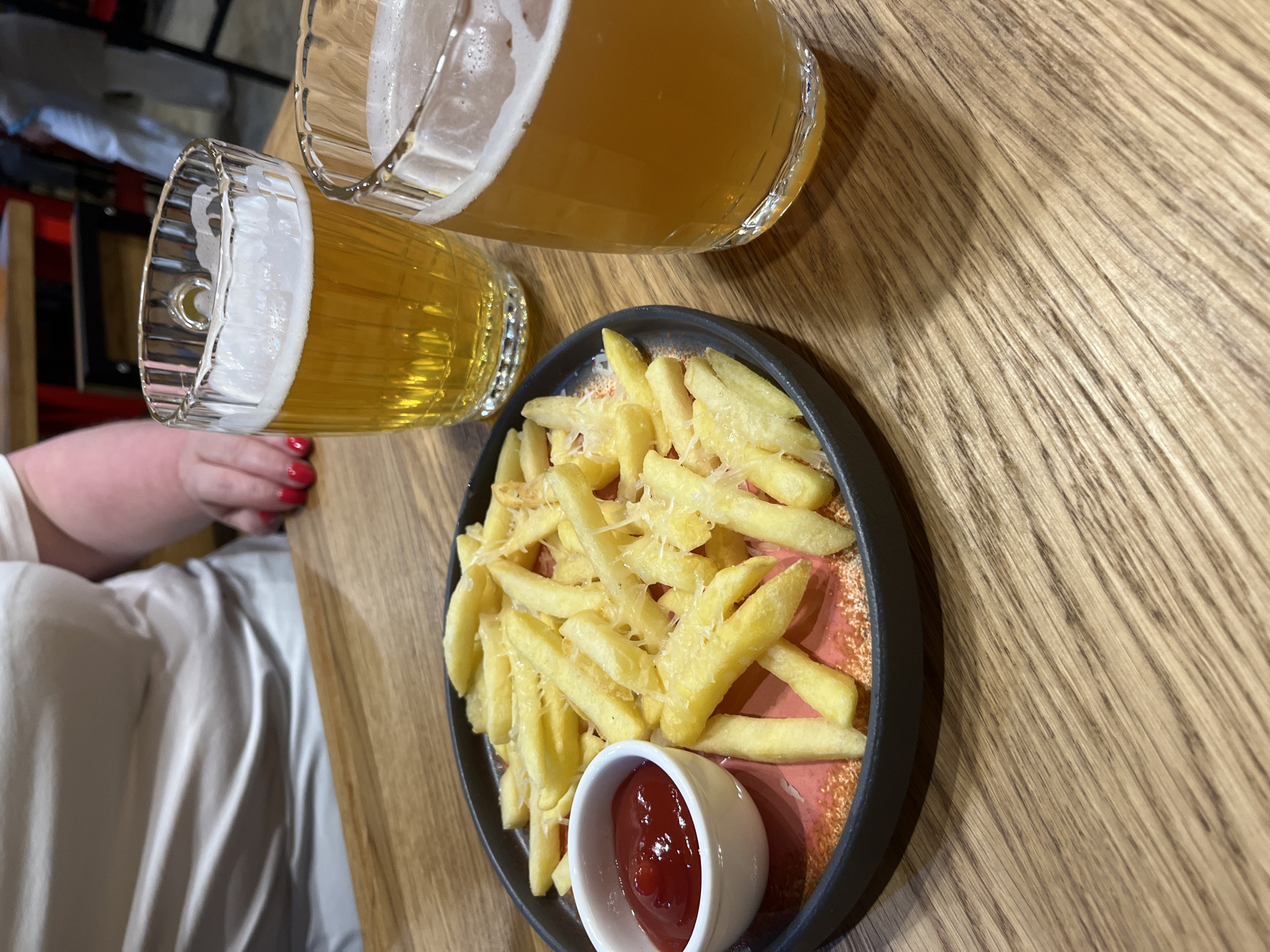 A close-up, high-angle shot shows a dark, circular plate of french fries sprinkled with cheese and a small white ramekin of ketchup, positioned on a wooden table. Two glasses of beer with frothy heads are also on the table, one in the foreground and one slightly behind and to the left. A person, whose torso and arms are visible from the chest down, is seated at the table, wearing a light-colored top. Their right hand, with red nail polish, rests near the plate. In the background, blurred figures and elements of a restaurant interior are visible, including a red chair and another person wearing white pants. The scene appears to be a casual meal or drinks at a restaurant in Ново-Никольское, Russia.