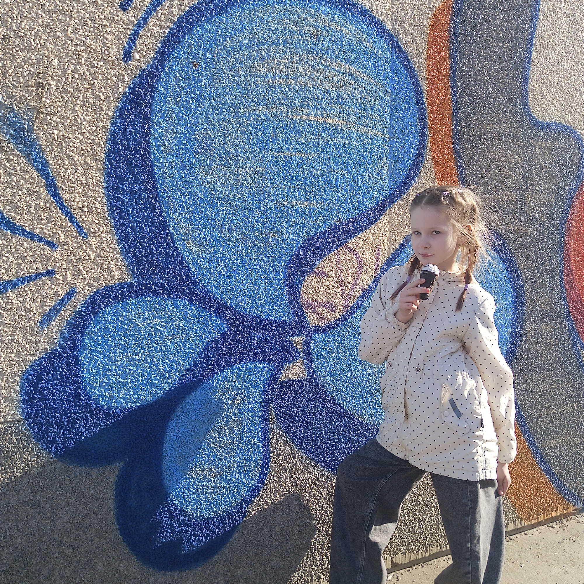 A young girl with braided hair stands against a wall with graffiti, holding an ice cream cone. The graffiti includes a large blue floral or abstract design. The girl wears a cream-colored jacket with black polka dots and dark jeans. The wall's texture is rough and granular. The location may be Chaikovsky, Russia, though there is no direct evidence of that in the image.