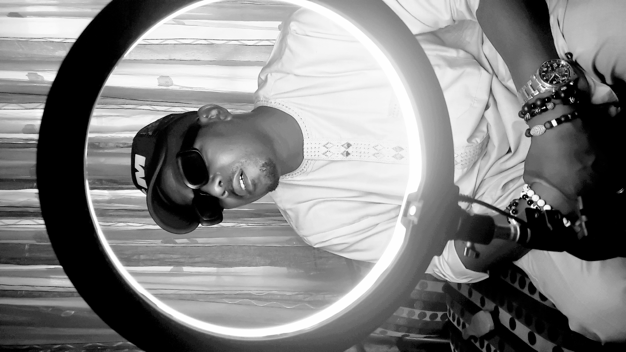 A black and white portrait shows a man wearing a baseball cap, sunglasses, and a light-colored, patterned shirt posing in front of a ring light. He also sports a watch and several bracelets on his wrist. The background features striped curtains. The man appears to be located in Narayi, Nigeria.
