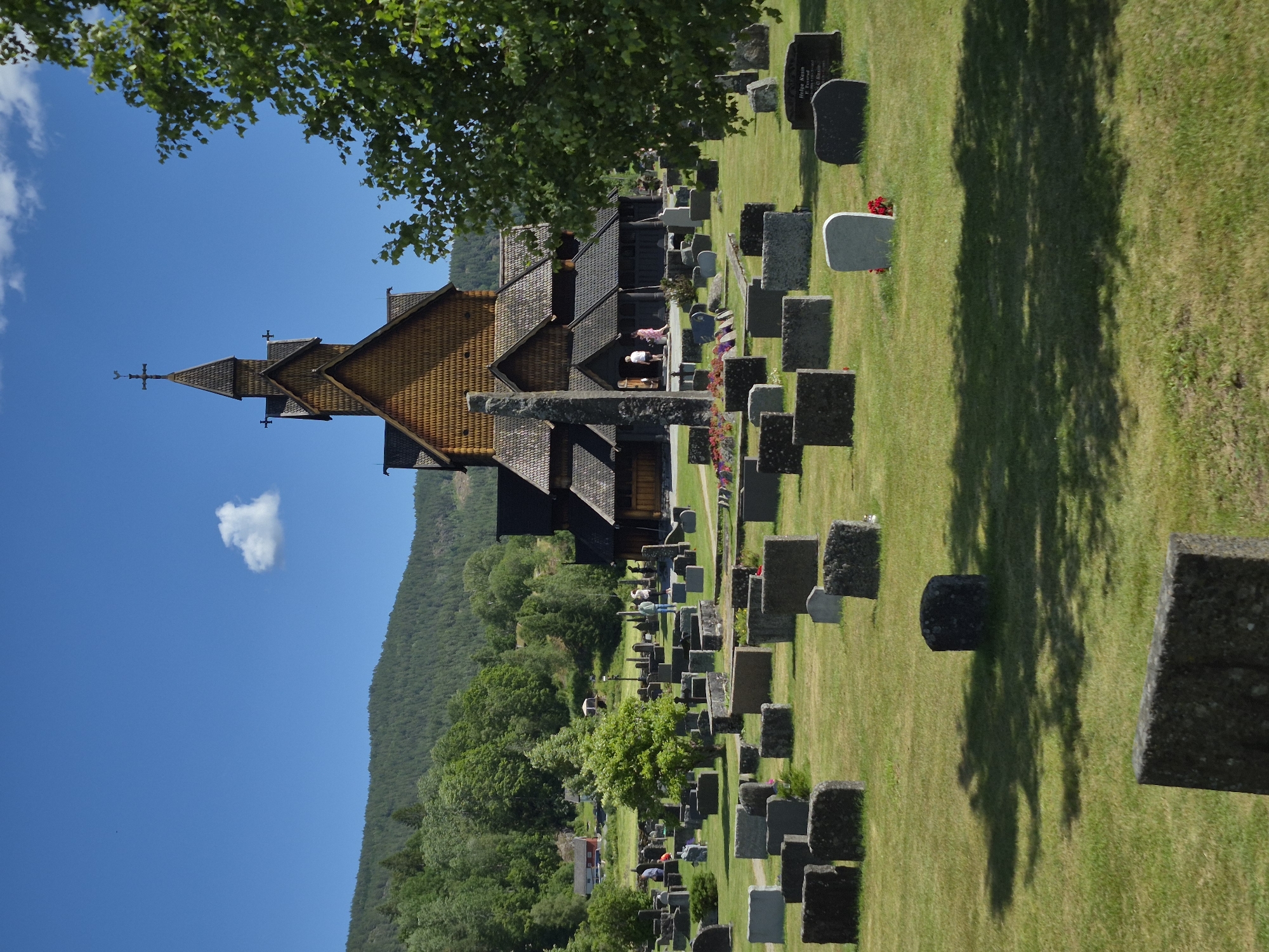This is a photograph of the Heddal Stave Church in Heddal, Norway. The wooden church, with its dark, shingled roof and steeple topped with a cross and weather vane, is the central focus. It is surrounded by a graveyard filled with gravestones of various shapes and sizes, many of which are dark and rectangular, while others are lighter and more rounded. A tall, stone monument stands in front of the church entrance. Several people are visible in the scene, including individuals standing near the church entrance and walking among the graves. The graveyard is set against a backdrop of lush green hills and a bright blue sky with a single white cloud. The sun casts shadows on the grass, indicating a sunny day. The environment is one of peace and solemnity, typical of a church and its surrounding burial ground.