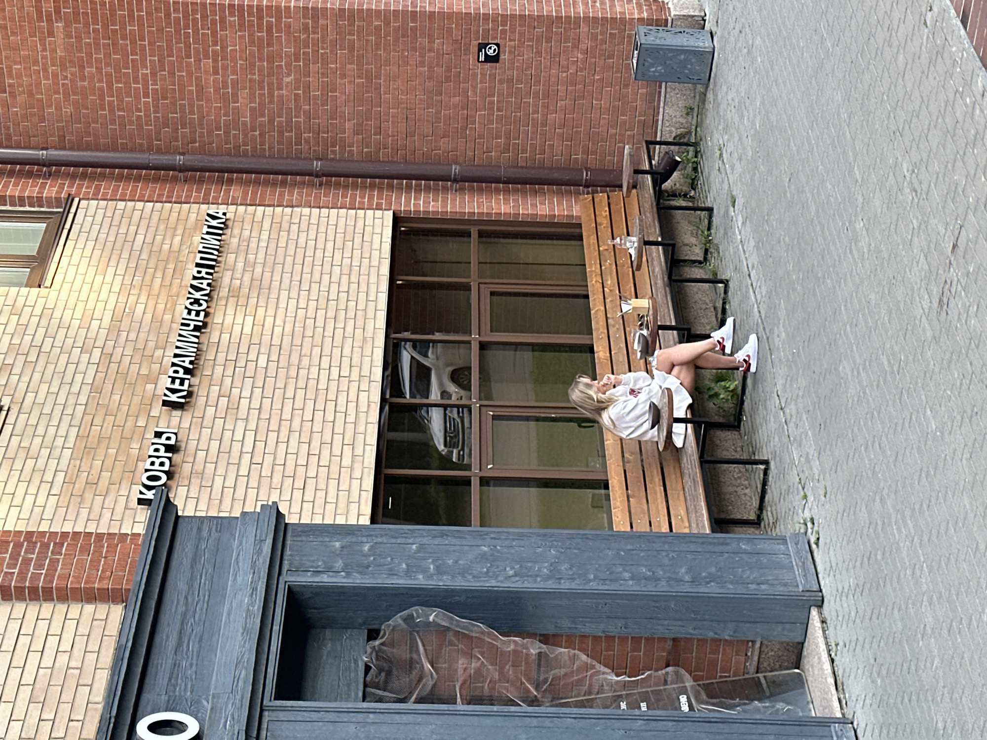 A woman is sitting on a wooden bench in front of a brick building. She is wearing a white sweatshirt and a white skirt, with white and red sneakers. The bench is situated next to a small table with drinks and some food items. The building has a sign with Russian text on it, which reads "КОВРЫ КЕРАМИЧЕСКАЯ ПЛИТКА". A dark-colored downspout runs down the side of the building. To the right of the bench, there is a metal trash can. In the background, through a window, a white van is visible. The ground in front of the bench is paved with gray bricks. The overall environment appears to be an urban setting.