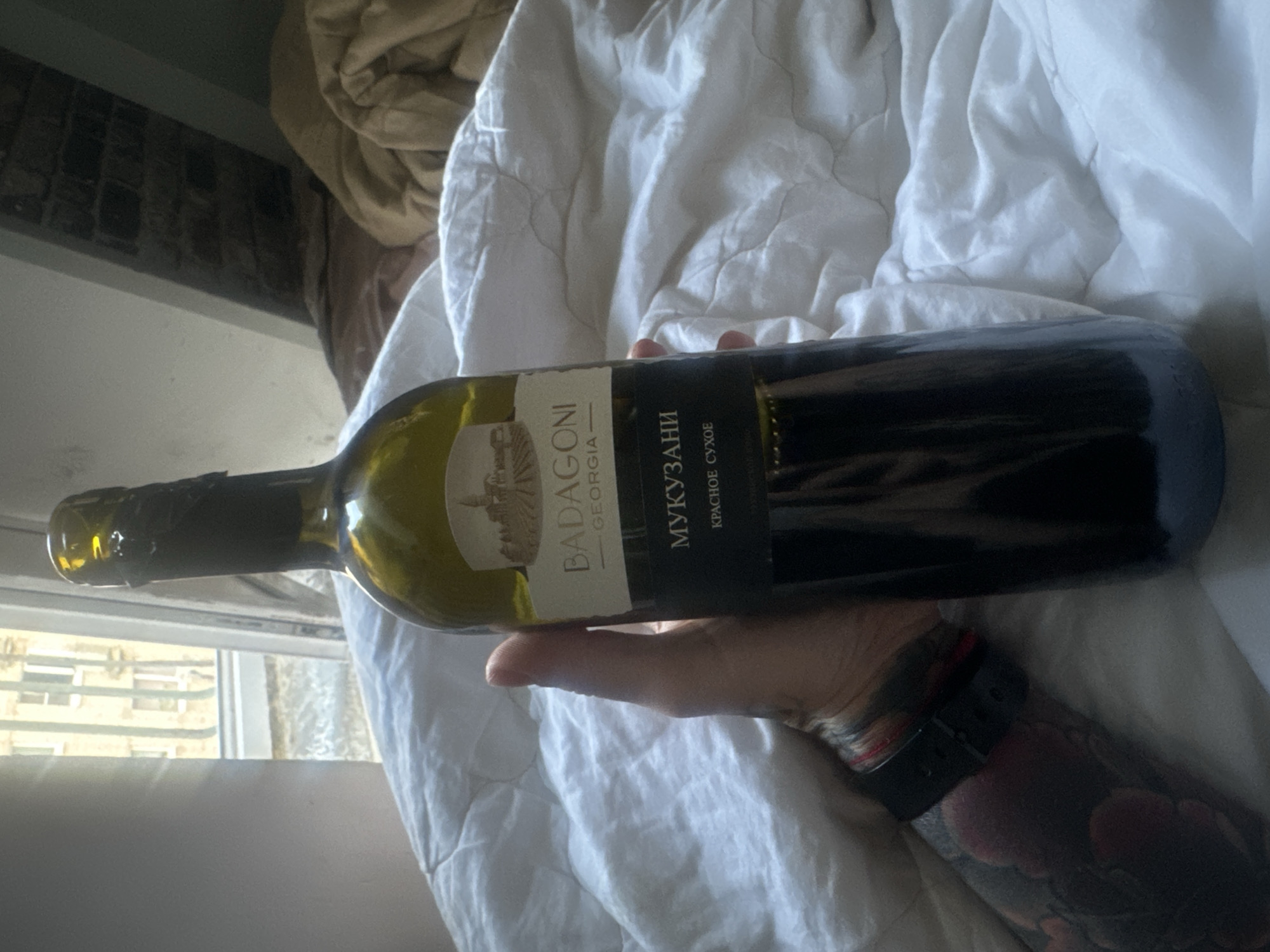 A hand with tattoos and a smartwatch is holding a bottle of Badagoni Mukuzani red wine on a white, quilted blanket. Light from a window with a cityscape is visible in the background. The location is unknown and cannot be identified as Chaikovsky, Russia.
