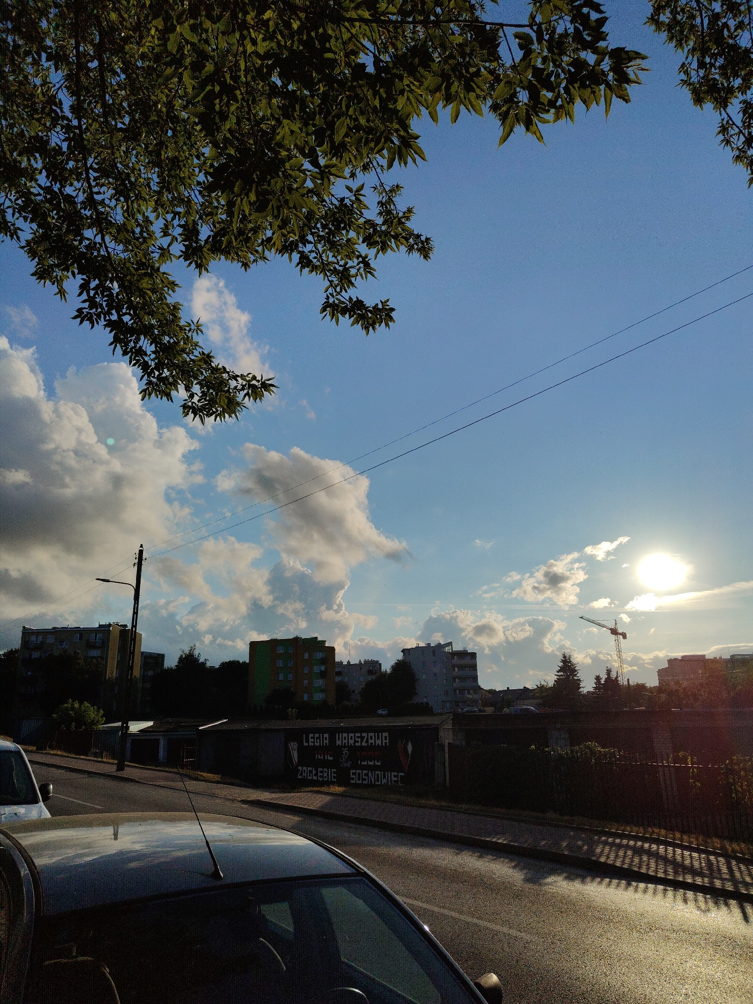 The image depicts an outdoor scene with a clear blue sky partially obscured by green tree foliage in the upper left and right corners. Several large, fluffy white clouds are scattered across the sky. In the distance, a city skyline is visible, featuring several multi-story buildings, some with a yellow or green facade. A prominent element in the background is a construction crane. A street runs diagonally from the lower left to the mid-right of the frame, with two cars visible on it. The foreground is dominated by the roof and windshield of a dark-colored car. A long, dark fence runs along the right side of the street, casting a patterned shadow on the asphalt. A street lamp stands on a tall pole on the left side of the image, and power lines are visible stretching across the sky. There is text on a wall behind the street lamp, which appears to be a mural or advertisement, reading "LEGIA WARSZAWA" and "ZAGŁĘBIE SOSNOWIEC". The sun is visible as a bright orb in the upper right quadrant of the sky, partially veiled by clouds, indicating it is either sunrise or sunset. The overall impression is an urban landscape bathed in the warm light of the setting or rising sun.