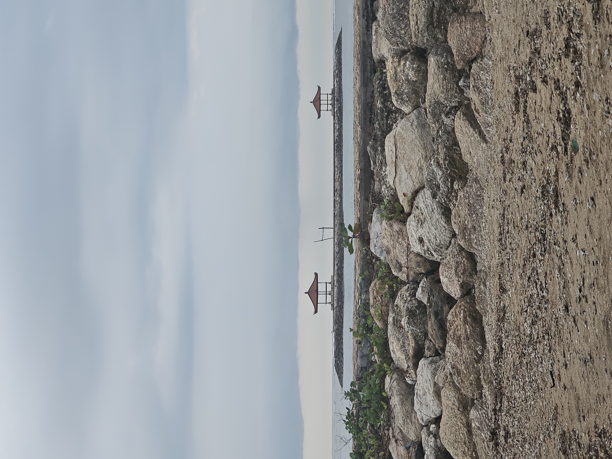 The image shows a coastal scene in Denpasar, Indonesia. The foreground features a stone wall covered in vegetation. Beyond the wall is the sea, with two small, red-roofed pavilions visible in the distance. The sky is overcast and light gray.
