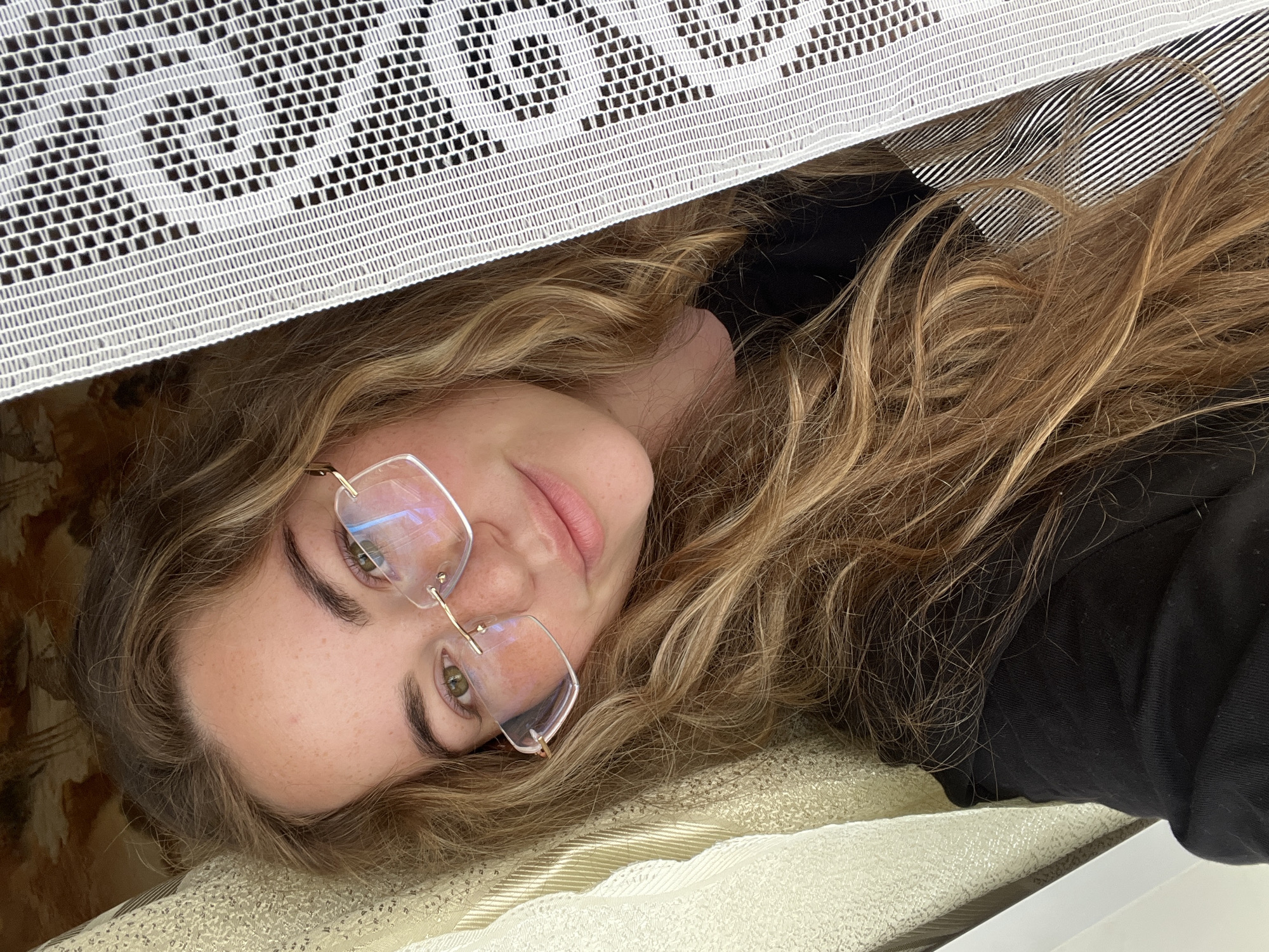 An indoor portrait taken in Сосново, Russia, captures a young adult female positioned horizontally, her head tilted to the left, gazing directly into the camera. She has long, wavy, light brown hair, fair skin with freckles across her nose and cheeks, and light green eyes. She wears rimless eyeglasses with thin gold-toned temples and a bridge. Her lips are slightly parted in a faint smile. She is dressed in a black crewneck top, and a garment with black and white vertical stripes is visible beneath a semi-translucent white lace fabric on her right side. This lace, characterized by a fine square grid pattern interspersed with larger dark square motifs, partially covers her right shoulder and torso, indicating it is likely a curtain or decorative textile. To her left, supporting or behind her head, is a light gold-colored fabric with subtle vertical stripes, consistent with a pillow or upholstered surface. The background directly behind her head and upper body appears to be a darker, ornate patterned fabric or wallpaper in tones of brown and gold. The scene suggests a relaxed, domestic environment.