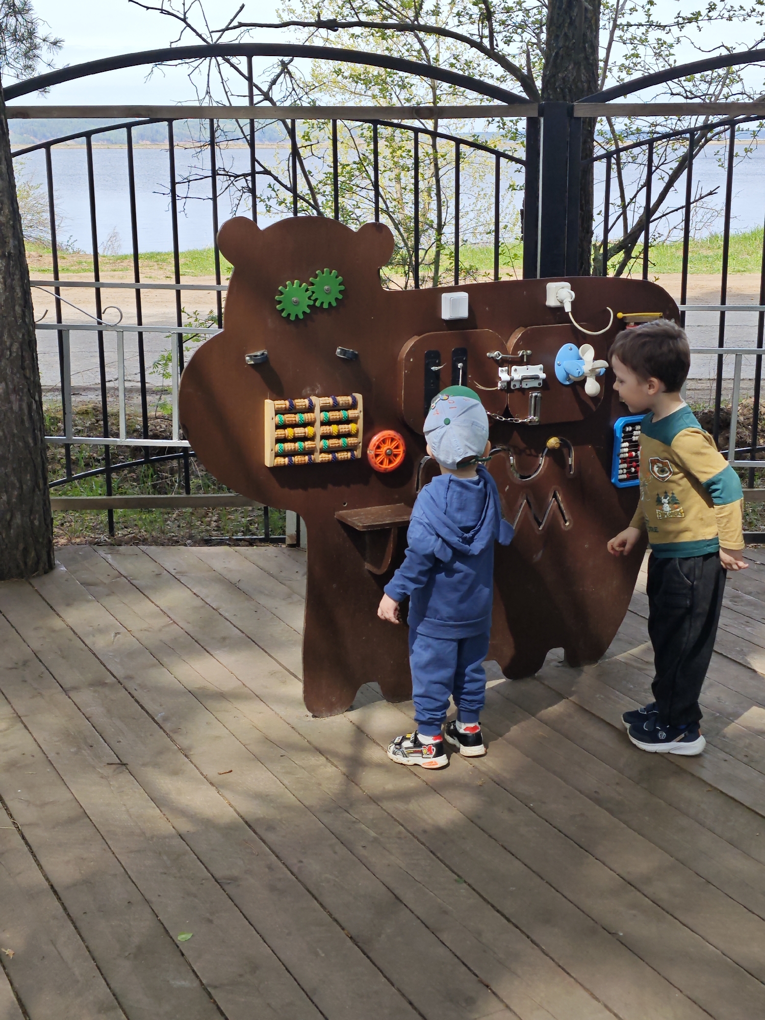 The image depicts two young boys interacting with a large, bear-shaped activity board on a wooden platform. The board is equipped with various manipulative elements such as gears, switches, and an abacus. The boy on the left is wearing a blue outfit and a hat, while the boy on the right is wearing a yellow and green shirt and dark pants. They appear to be engaged in playing with the board's features.
Behind the platform, a metal fence separates the area from a view of a body of water and some greenery. The presence of trees and the type of fencing suggests that this is an outdoor playground or recreational area.
The location is identified as the city of Chaikovsky, Russia, based on visual cues such as the architecture and environment.