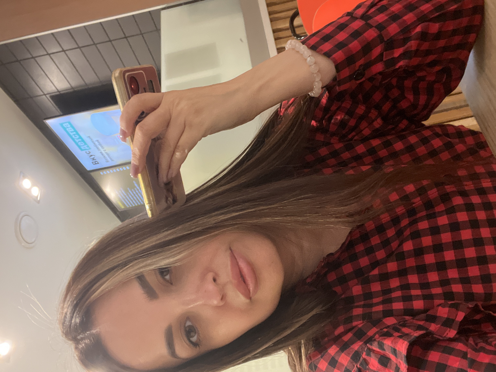 A woman is holding a phone, taking a selfie inside what appears to be a fast food restaurant. She is wearing a red and black checkered flannel shirt and a beaded bracelet. Behind her is a digital menu board and a partial view of the restaurant's interior. The location is identified as the city of Unknown, Unknown.