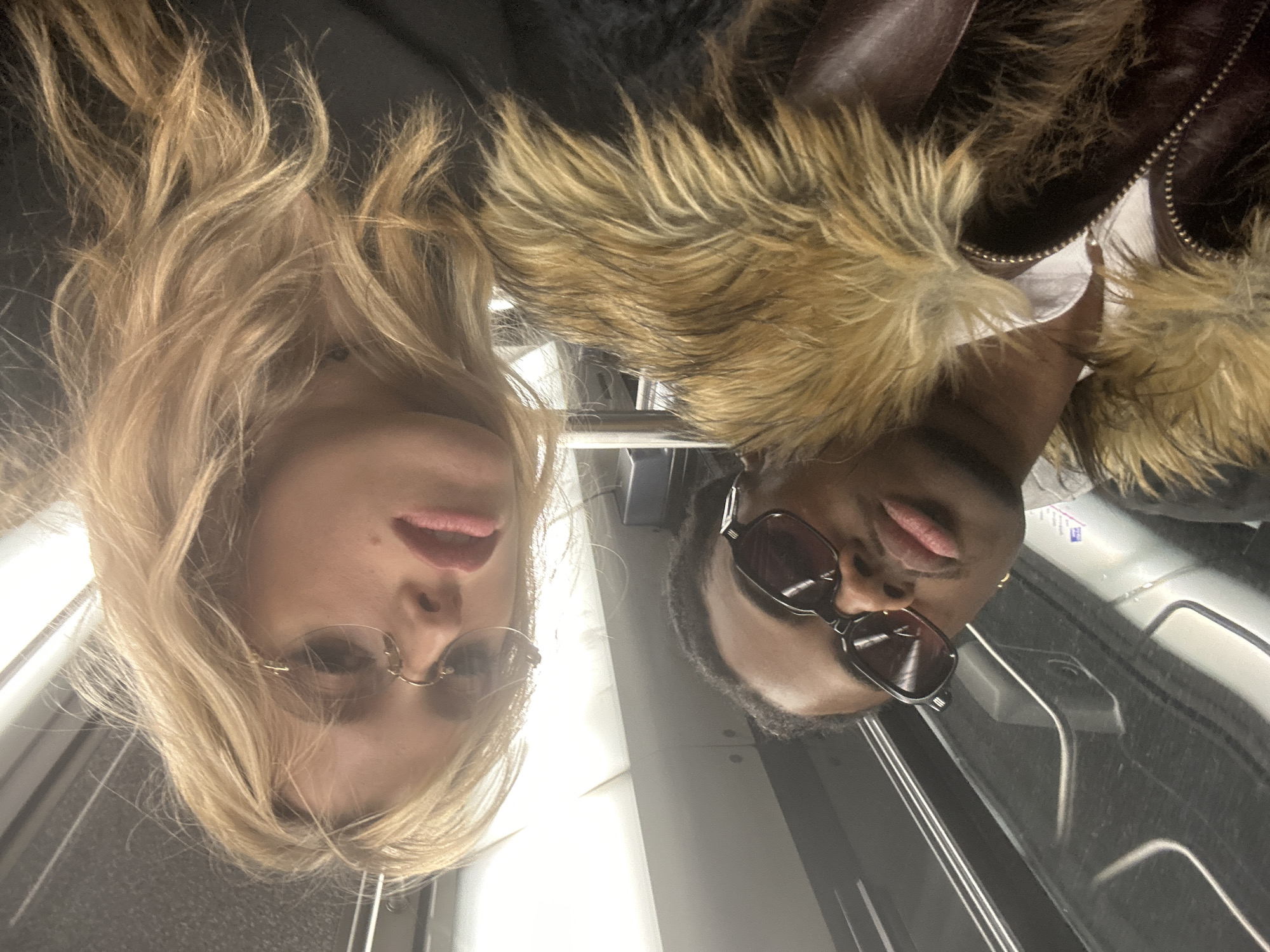 Two people, one fair-skinned with blonde hair and one dark-skinned with short black hair, are on a Paris Metro train. The fair-skinned person wears rimless sunglasses. The dark-skinned person wears dark framed sunglasses and a brown leather jacket with a fur-lined collar. The image is taken from a low angle, looking up. The interior of the train is visible, including the seats, windows, and handrails. The location is likely a station or inside of the metro train in Paris, France.