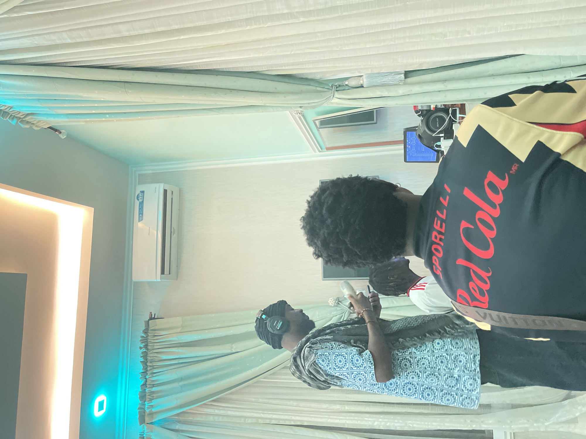 Three men are present in a room, likely in the city of Gura Top, Nigeria. One man is wearing headphones and a patterned scarf. Another man stands with his back to the camera, wearing a black shirt with "Red Cola" in red lettering. A third man is visible between them. The room has white walls, white curtains, and a bright light source on the left. An air conditioner is mounted on the wall. A camera is present on a tripod.