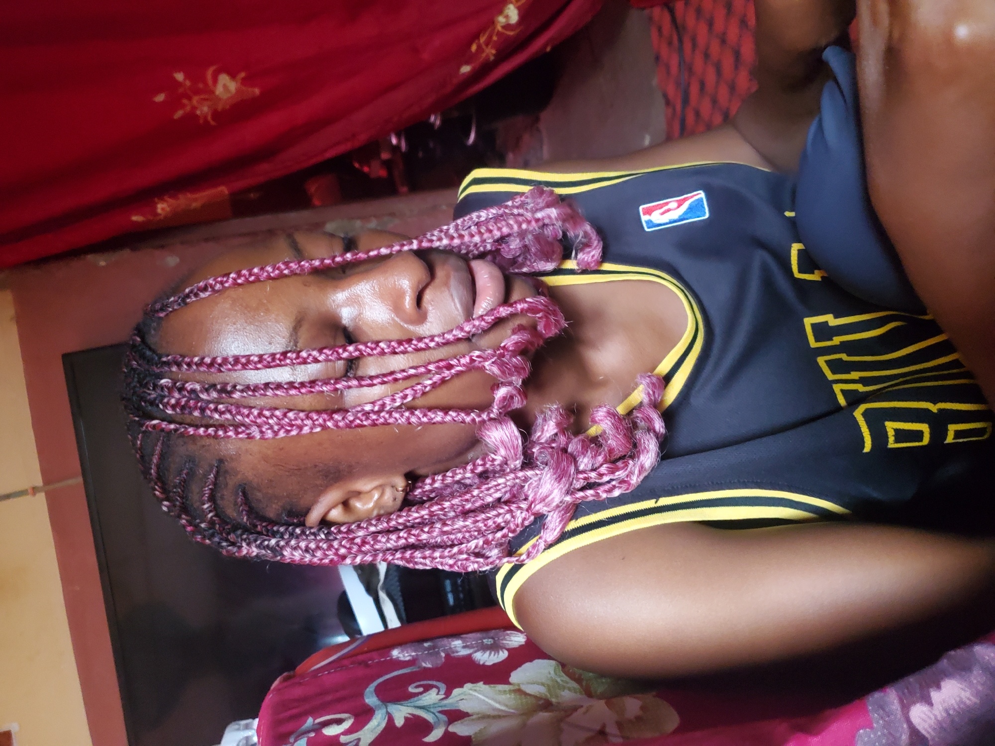 The image shows a person lying down, possibly asleep, with their eyes closed. The person has braids in their hair, with some strands of pink. They are wearing a black and yellow athletic jersey with the NBA logo on the left side of the chest. The background includes red fabric with gold patterns and a glimpse of a red and white floral patterned cloth. The scene appears to be indoors, with warm lighting. The overall mood is one of rest and relaxation. There is no visible text other than the NBA logo. The location context provided is Jos, Nigeria.