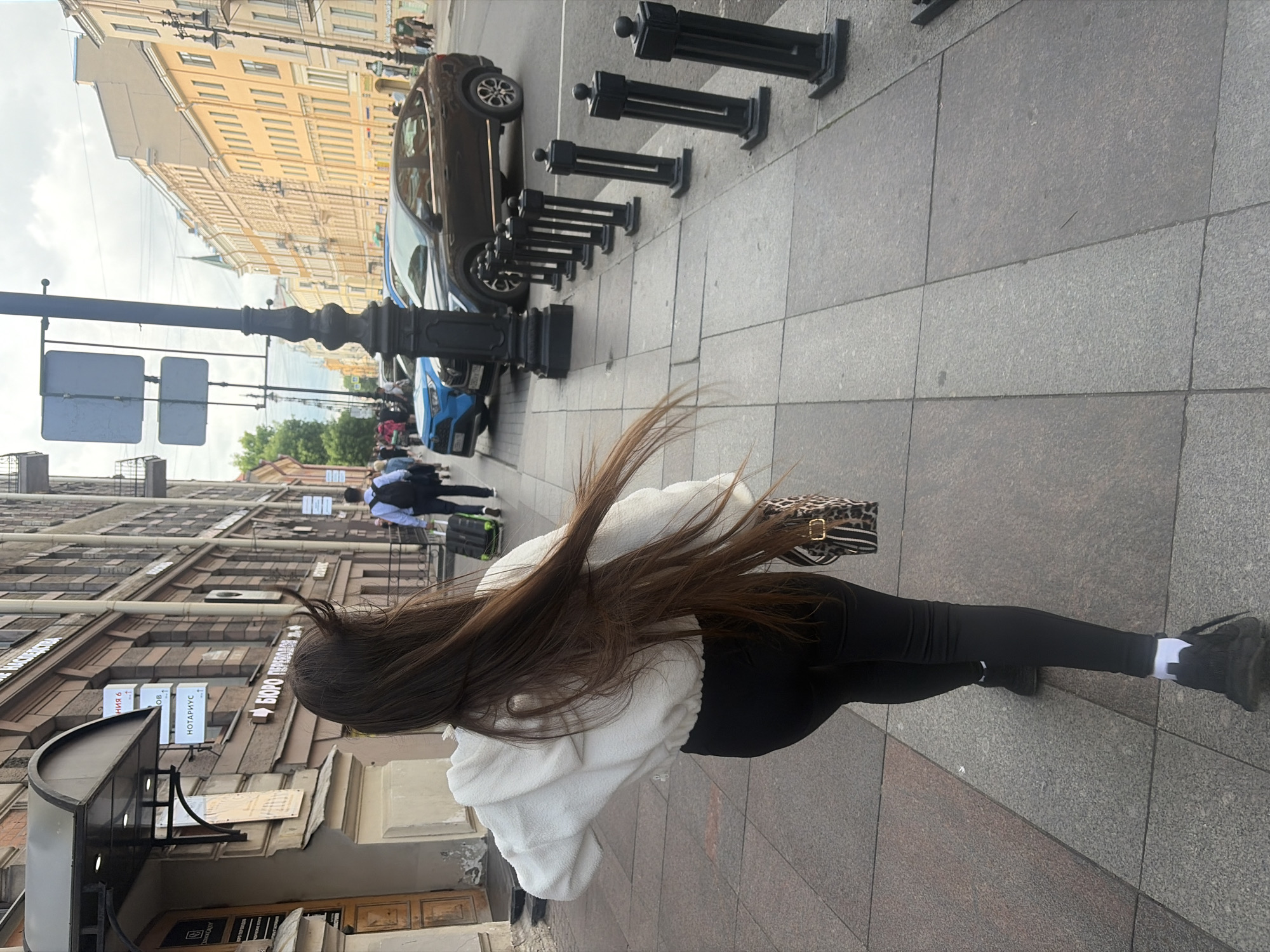The image depicts a street scene in Saint Petersburg, Russia, likely on Nevsky Prospekt or a similar major avenue. A young woman with long, dark hair is walking away from the camera on a wide sidewalk paved with large grey granite slabs. She is wearing a white fuzzy hooded jacket, black leggings, white socks, and black athletic shoes. She is carrying a small handbag with a leopard print design and a black and white strap. Her hair is being blown to her right by a strong wind.
In the background, other pedestrians are visible, including a man in a blue shirt and black pants pulling a dark green suitcase on wheels. Several cars are parked along the street, including a dark brown crossover SUV and a bright blue sedan. The buildings lining the street are typical of Saint Petersburg's architecture, with multiple stories, ornate facades, and large windows. Signs in Russian are visible on some of the buildings, including one that reads "БЮРО ПЕРЕВОДОВ" (Translation Bureau). A black lamppost with ornamental detailing stands on the sidewalk in the foreground. The sky is overcast with a mix of grey clouds.