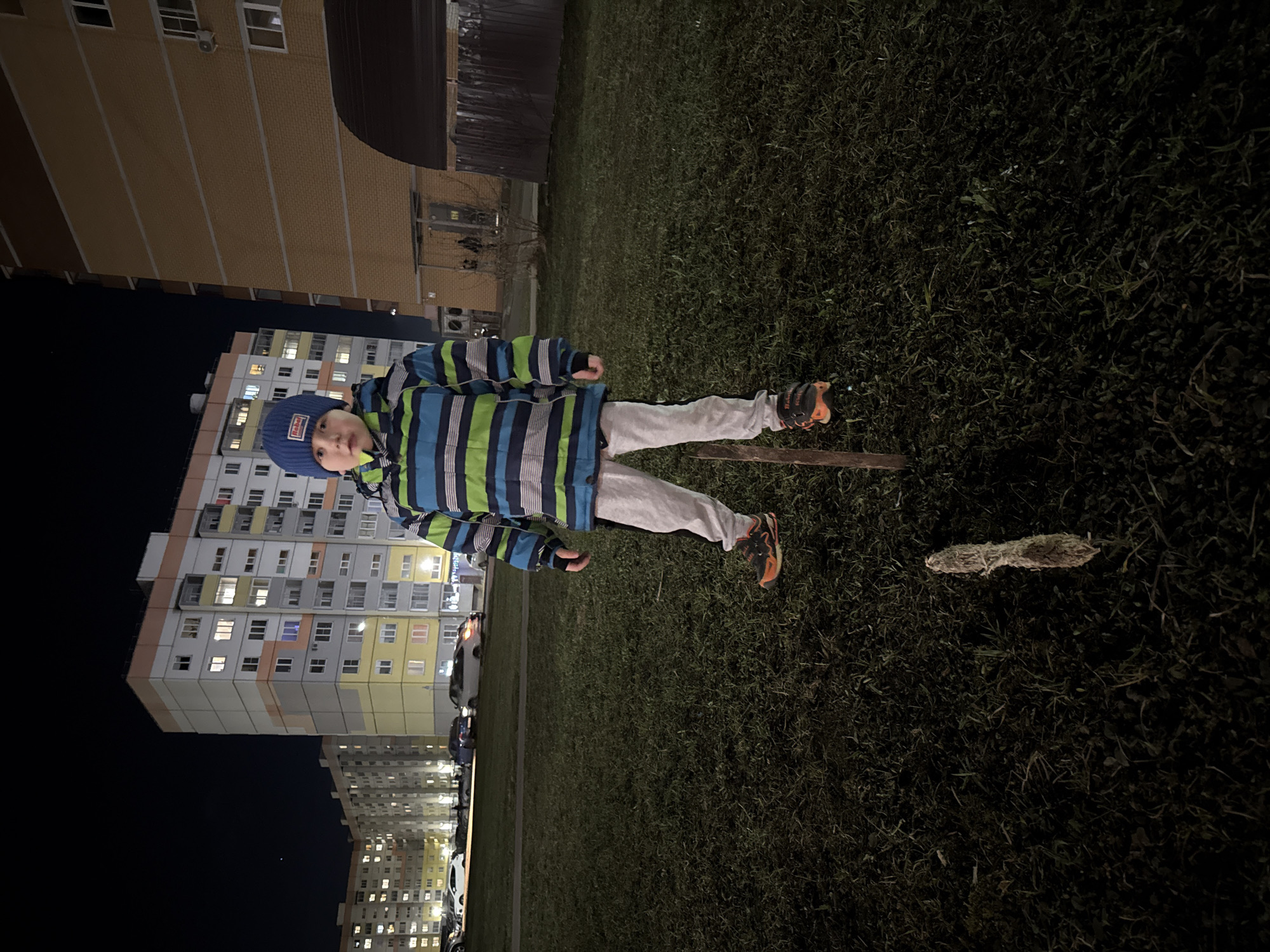 The image depicts a child standing on a grassy area in an urban environment at night.
The child is wearing a blue hat with text, a striped blue, green, and white jacket, gray pants, and orange and black sneakers. They are standing on grass, near a small stick or branch. The ground appears to be grassy. Behind the child is an apartment building. The presence of multiple residential buildings suggests the setting is likely a residential area in a city. Veliky Novgorod, Russia is a possible location.