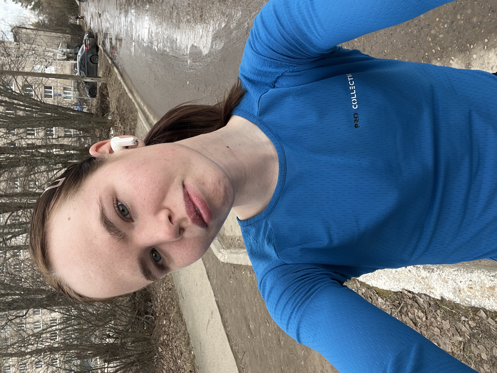 A woman with light skin and brown hair, wearing a blue long-sleeved shirt, is taking a selfie outdoors. She has an earbud in her left ear. The background shows a street scene in Chaikovsky, Russia. Bare trees line the left side of the frame, and a multi-story building is visible behind them. The street is wet, with melting snow or ice in some areas. A dark car is parked along the side of the road. The woman is standing near a curb, with fallen leaves and debris on the ground.