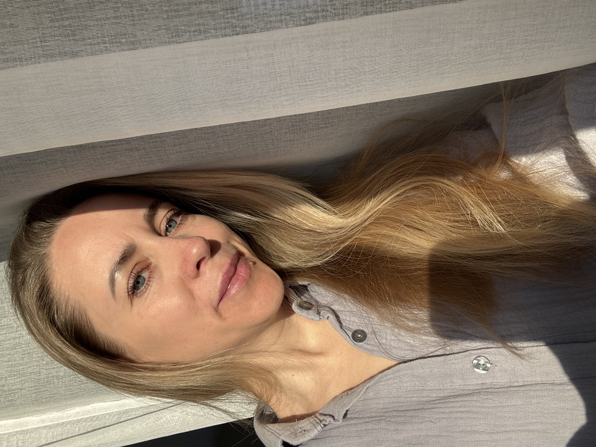 A fair-skinned woman with blonde hair is lying down, possibly in a bed, as suggested by the visible fabric. She is wearing a light grey button-up shirt. Light from a window illuminates her face and the top of her shirt, casting shadows. The context is unclear, and there is no information available in the image to place it specifically in the city of Chaikovsky, Russia.