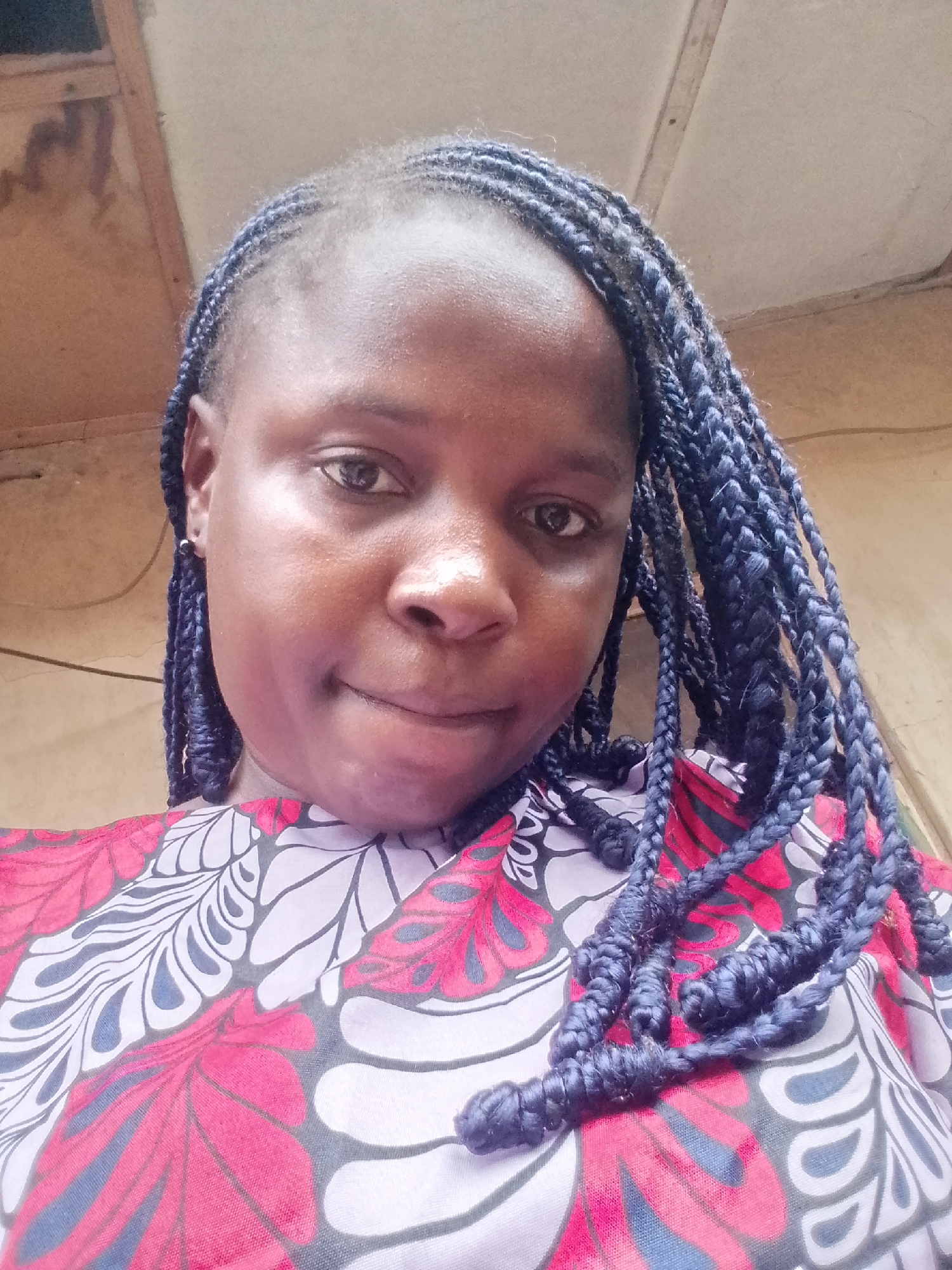 A selfie of a dark-skinned woman with dark braids adorned with blue highlights, wearing a vibrant patterned top with red and white floral designs. The background is blurry and indistinct, appearing to be an interior space with light-colored walls and a wooden ceiling. The woman is gazing directly at the camera with a slight smile. The image gives no information to guess the city Unknown, Unknown.