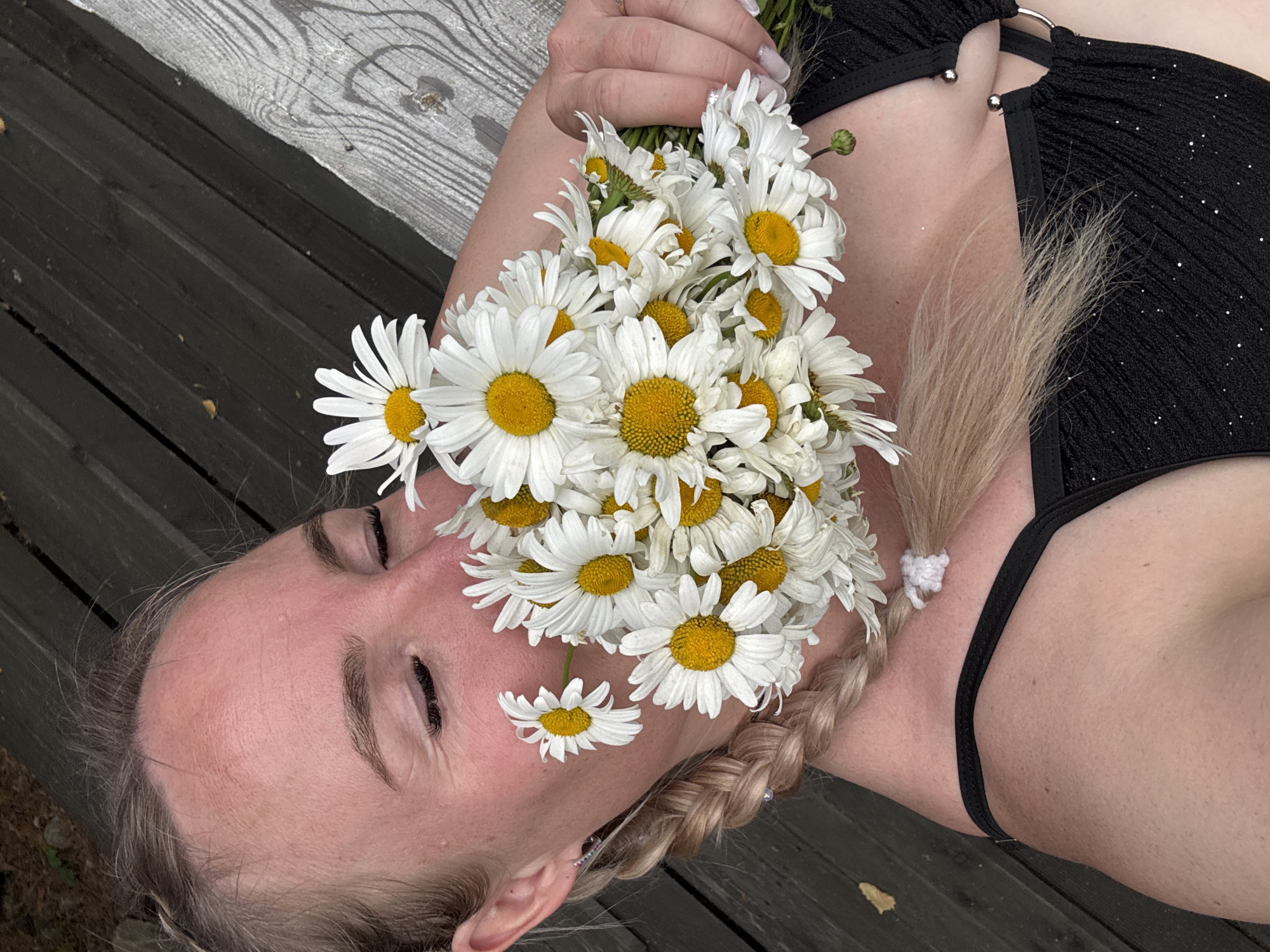 A person lies on a wooden surface, with a bouquet of daisies arranged around their head. The daisies are white with yellow centers, and some are placed directly on the person's face. The person's eyes are closed, and their expression is peaceful. Their hair is blonde and braided. They are wearing a black, sparkly bikini top. The wooden surface appears to be part of an outdoor seating area, possibly a bench or deck. The scene is set in Pozdyshki, Russia. The lighting suggests it could be daytime, with natural light illuminating the subject. The overall mood is serene and idyllic.