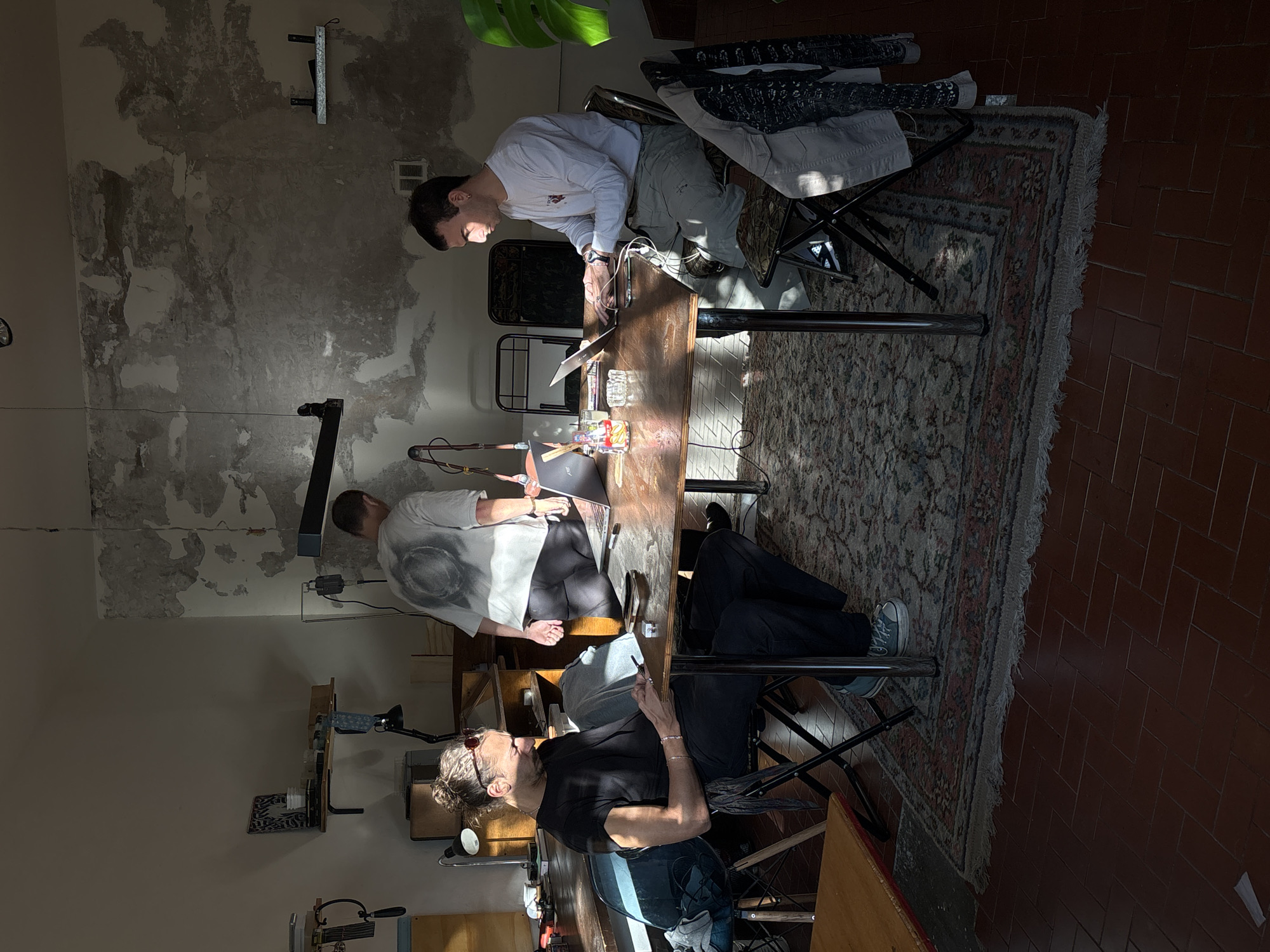 The image captures an indoor scene, likely a workshop or studio, in Buenos Aires, Argentina, illuminated by strong, direct sunlight. The lighting creates distinct patterns of light and shadow across the space.
Three individuals are present, gathered around a long, rectangular wooden table. In the foreground, a woman with her hair tied up and sunglasses resting on her head, dressed in a black t-shirt and dark pants, is seated at a folding chair. She is looking towards the far end of the table.
On the opposite side of the table, slightly further back, a man in a white long-sleeve shirt and light-colored pants is seated, deeply engrossed in working on a laptop. His hands are on the keyboard, and his face is illuminated by the sunlight. Behind him, draped over another folding chair, are various clothes or fabrics.
Further along the table, away from the foreground woman, sits another man wearing a white t-shirt with a large grey circular design. He is leaning over the table, intently focused on smaller items arranged there, suggesting detailed work.
The wooden table itself holds several objects: at least two laptops, a glass containing a drink with ice and a lemon wedge, a clear glass ashtray with a cigarette, and an assortment of small components or tools. The floor beneath the table features a patterned Persian-style rug, which transitions to reddish-brown terracotta tiles. The walls of the room have a textured, worn appearance, with areas of peeling paint and exposed plaster, lending a rustic or industrial character to the setting. A vibrant green plant leaf is visible in the upper right portion of the frame.
The scene suggests a focused, collaborative work environment during a sunny daytime, with no explicit text visible.