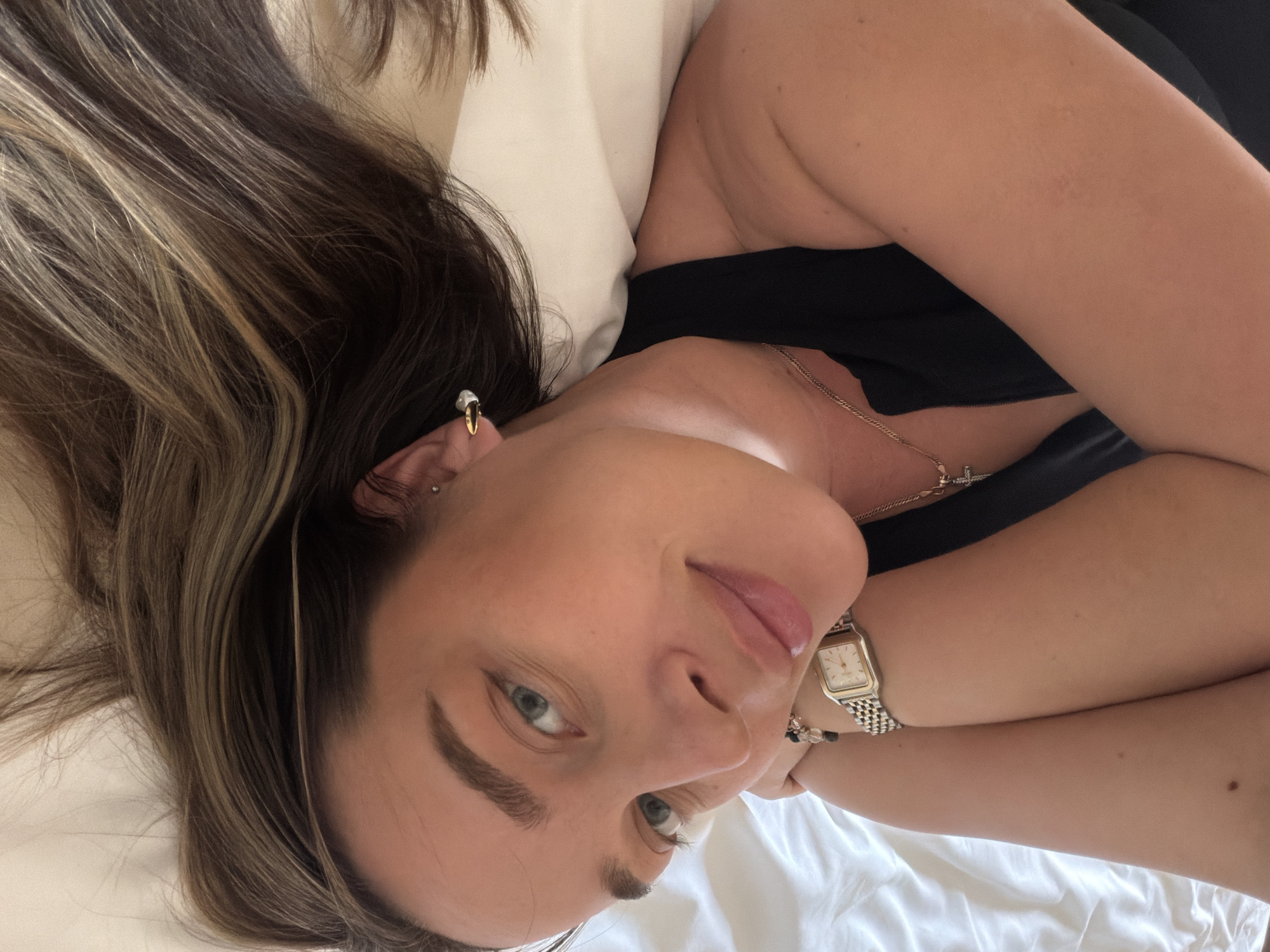 A woman with long, brown hair with blonde highlights, wearing a black top and a gold necklace with a cross pendant, is lying on a white bed with a white pillow. She is wearing a gold watch and bracelets on her right wrist, and she has earrings in her ear. The scene does not appear to be in Moscow, Russia.