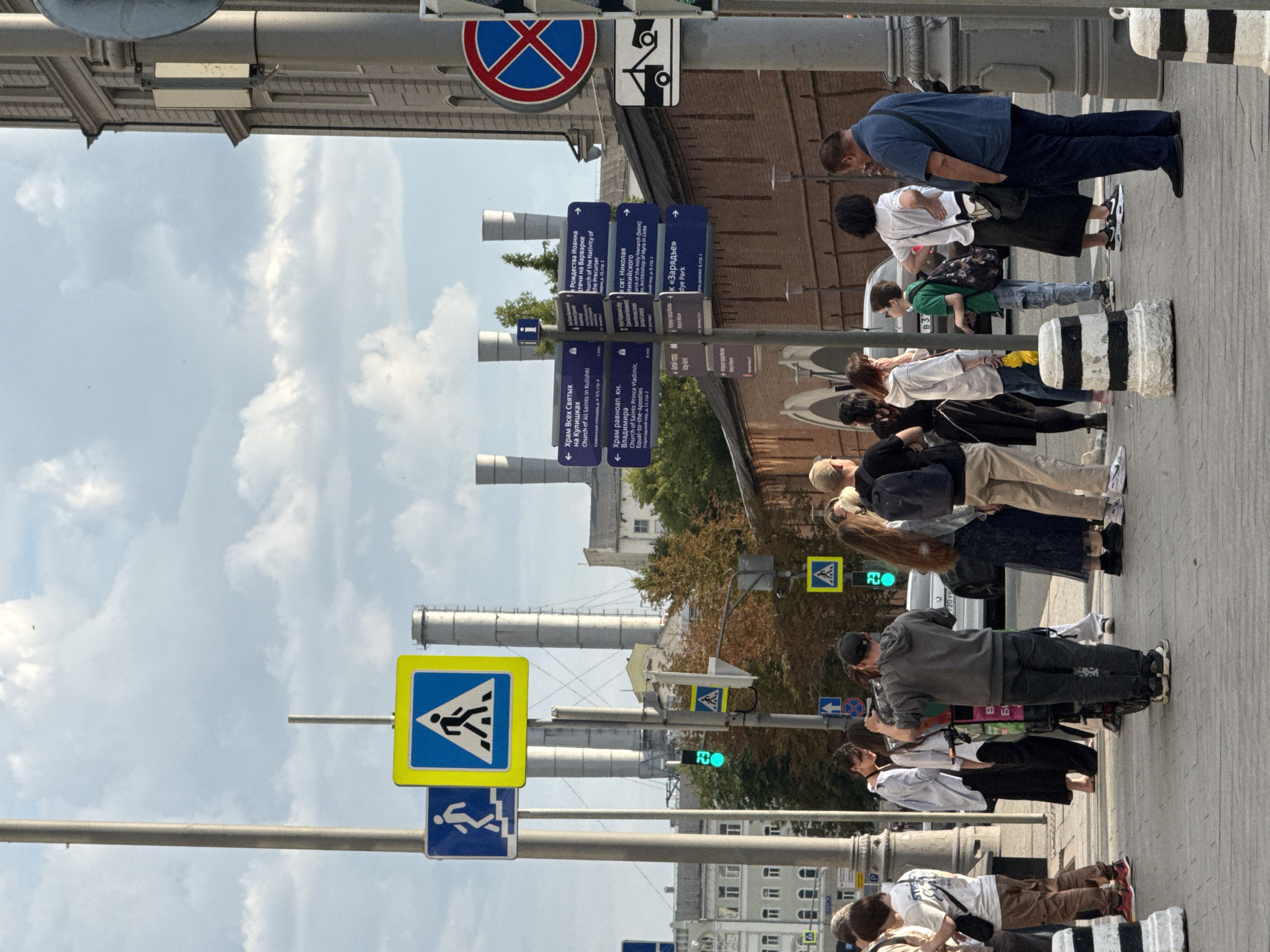 The image captures a street scene in Moscow, Russia, with a group of people standing on a sidewalk near a building with classical architecture and industrial chimneys in the background. Several directional signs are visible, indicating various churches and places in the city, such as "Church of All Saints in Kulishki," "Church of Saint Vladimir, Equal-to-the-Apostles," "Church of the Nativity of the Virgin Mary," and "Dye Park." A pedestrian crossing sign is prominently displayed, along with a "no stopping" sign. The sky is partly cloudy. The people in the image are casually dressed and appear to be tourists or locals. Some are looking at their phones, while others are observing their surroundings. A man in a grey hoodie is operating a camera on a stand, suggesting a filming or photography activity.