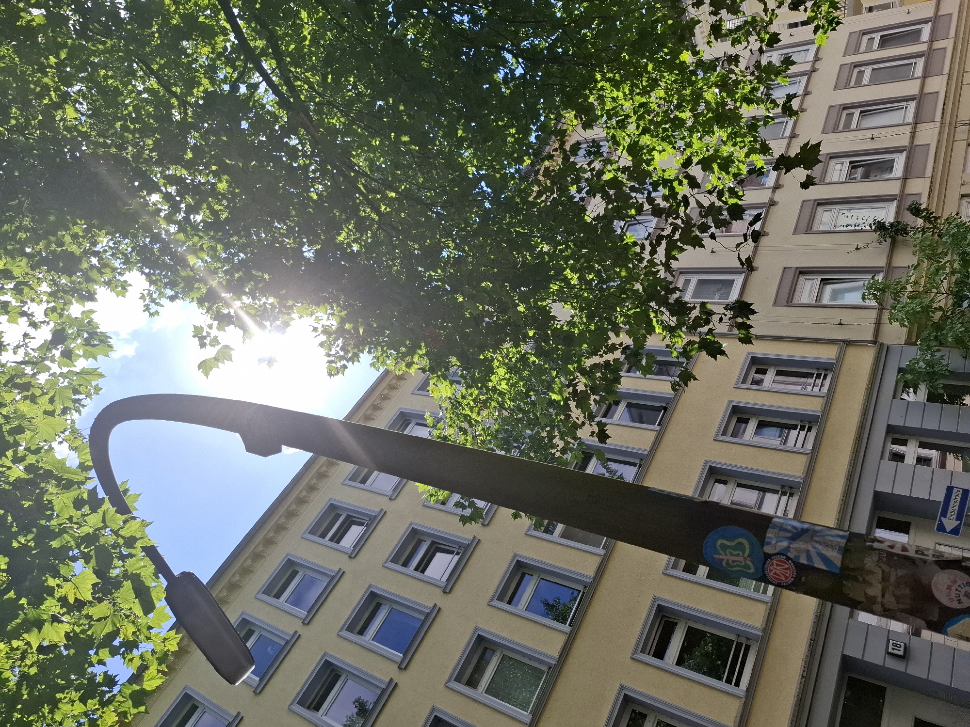 The image depicts a street scene in Hamburg, Germany, captured from a low angle looking upwards. A tall, curved street lamp with a dark finish dominates the left side of the frame. The lamp post is adorned with several stickers, one of which features a butterfly graphic and another with German text. Behind the lamp, a multi-story yellow building with numerous windows is visible. The windows have light-colored frames and many appear to have shutters. The top of the building features decorative molding. Lush green leaves from a large tree canopy the upper portion of the image, with sunlight filtering through them, creating dappled light and lens flares. The sky is blue with scattered white clouds. In the lower left corner of the building's facade, a house number "18" is visible. A blue sign with white text indicating "Einbahnstraße" (one-way street) is mounted on a pole near the bottom of the frame. The scene suggests a sunny day in a residential or mixed-use urban area.