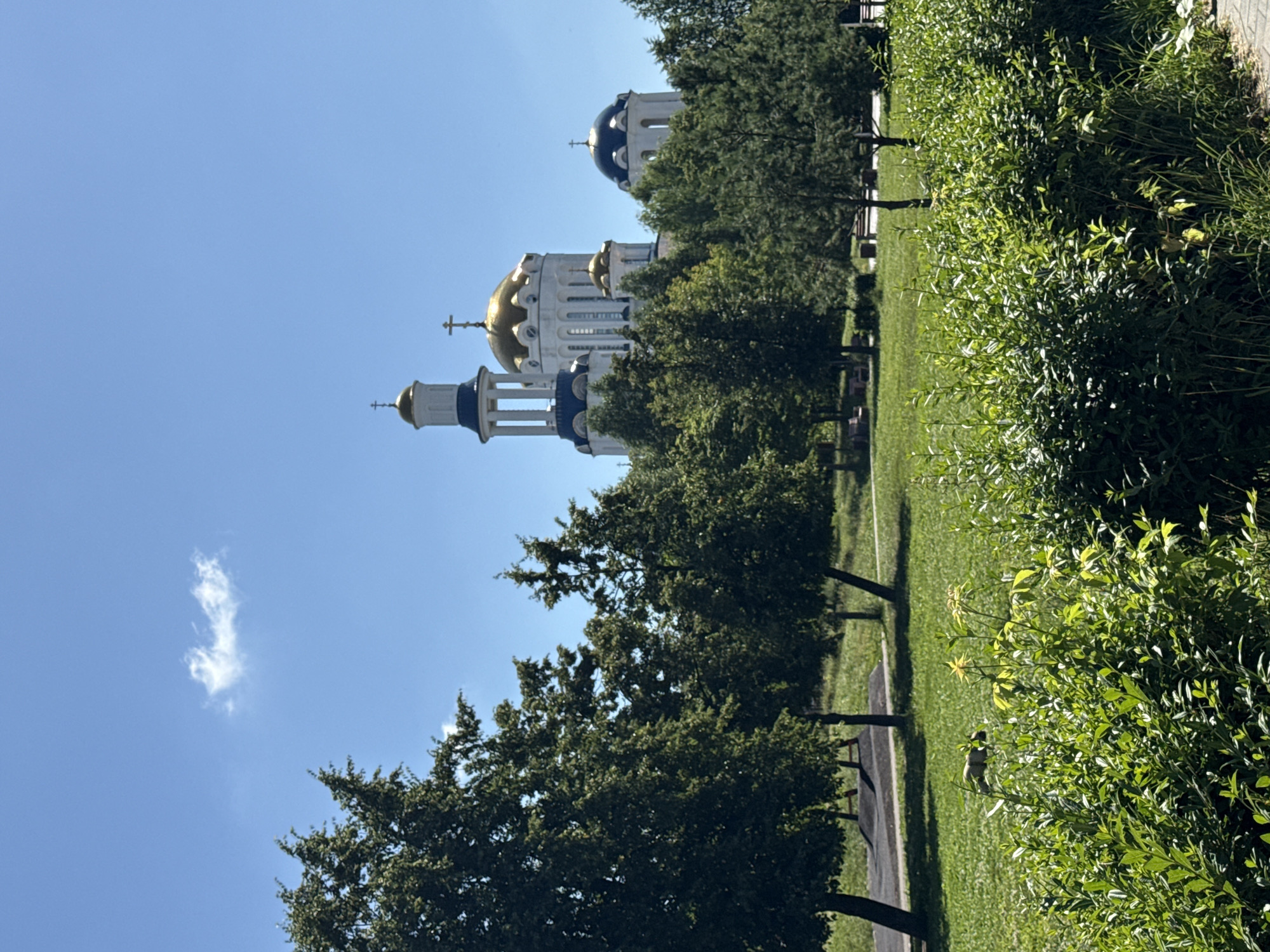 This is a vertical, full shot of a park with a large church in the background under a clear blue sky with a single wispy cloud. The church has multiple domes, the main dome is gold, and others are blue. The church has white walls and is surrounded by trees. The park area in the foreground is covered with lush green grass and dense bushes. There are some trees in the middle ground and playground equipment visible. The church is likely The Church of the Resurrection of Christ in Krylatskoye, Moscow, Russia.